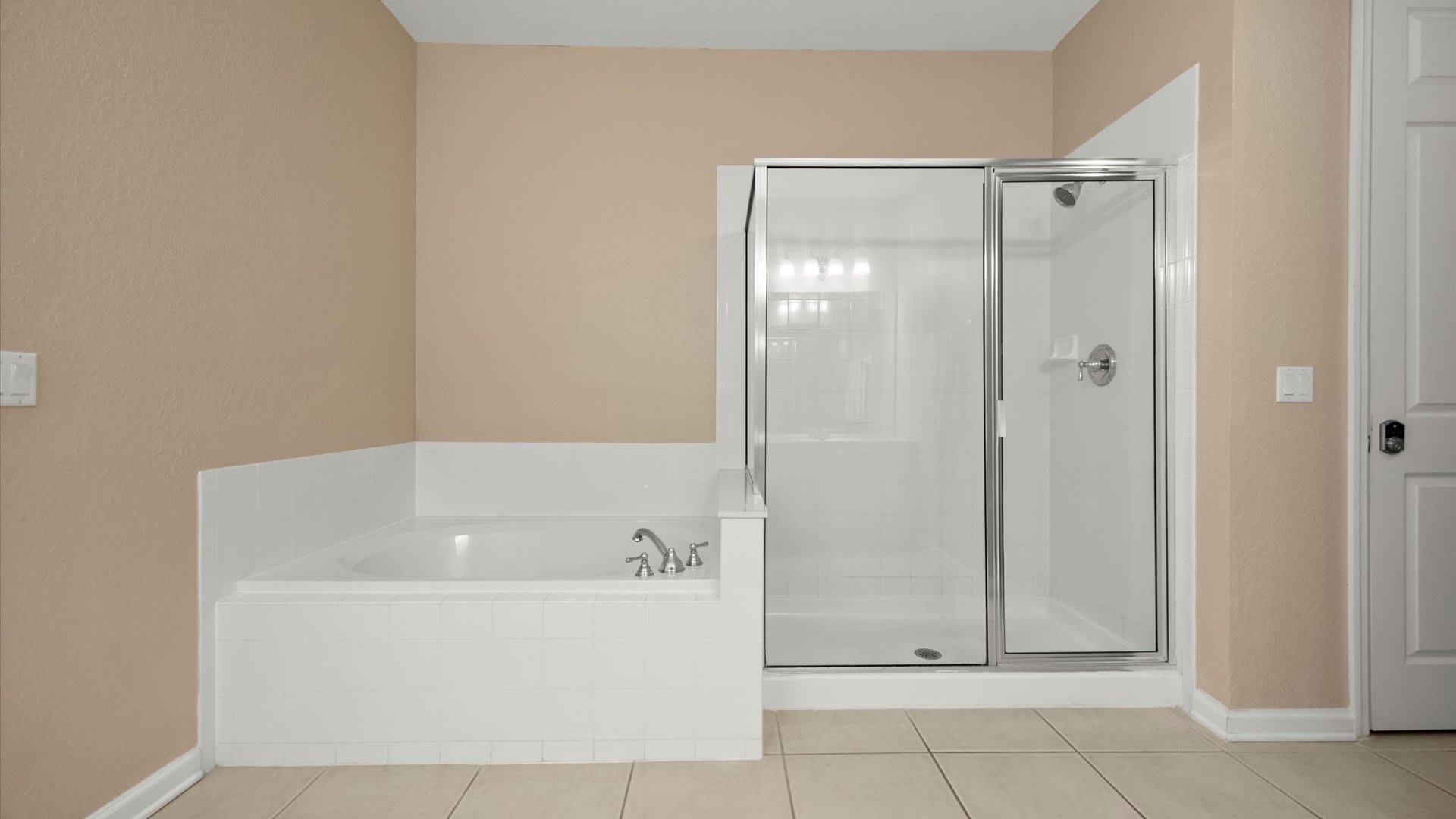 Close-up view of a white built-in bathtub with chrome fixtures and a glass-enclosed shower with white tiled walls in a spacious bathroom. The light neutral tiled floor complements the beige wall color.