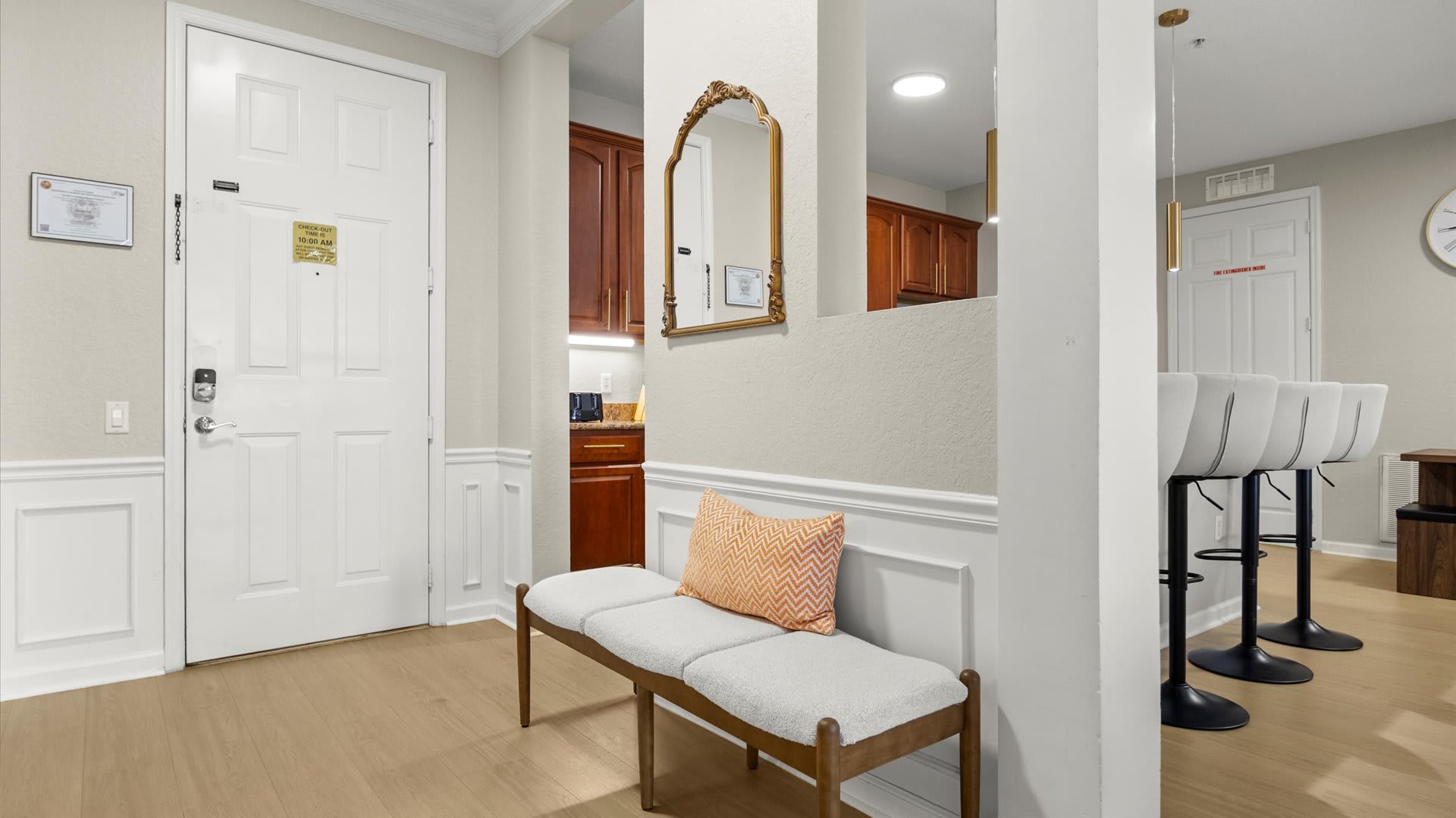A bright foyer with light wood flooring features a white paneled entry door with a digital lock, a light gray upholstered bench with a patterned orange pillow, and a gold-framed ornate mirror above wainscoting. A glimpse of a kitchen with dark wood cabinets is visible through an archway.