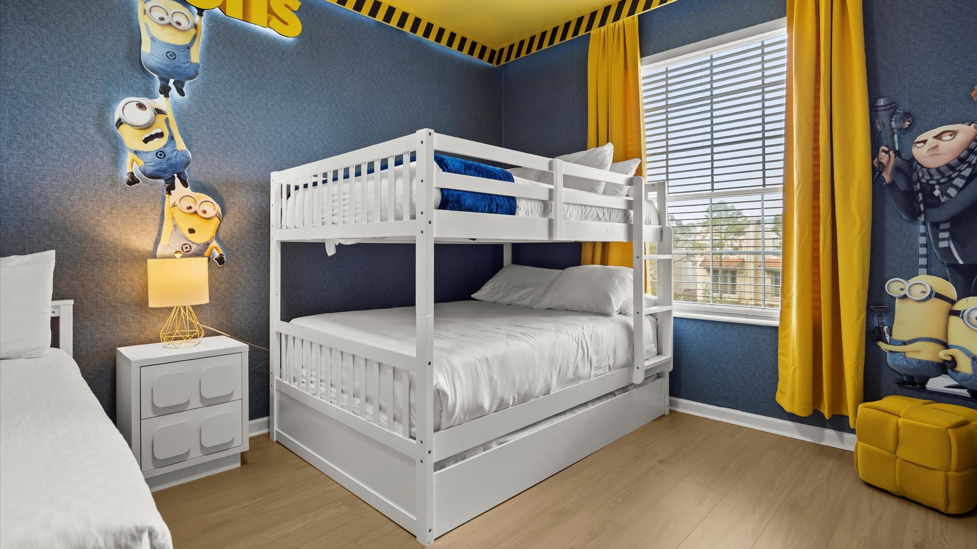 Minions-themed kids' bedroom with a white bunk bed (full over full with trundle), a full-size bed, and a window with yellow curtains. The room has blue walls and a yellow ceiling, all on a light wood floor.