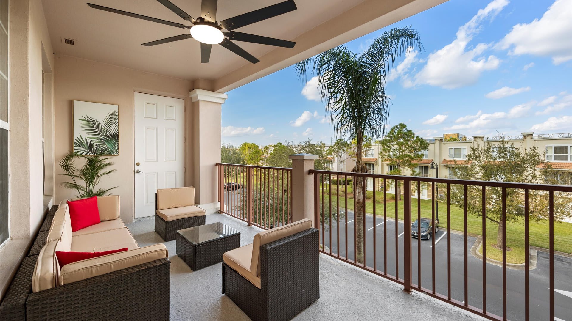 A spacious outdoor balcony furnished with a wicker sofa, two armchairs, and a coffee table, all with beige cushions. A ceiling fan provides comfort, and the balcony offers views of a parking area and surrounding buildings, with a palm tree visible.