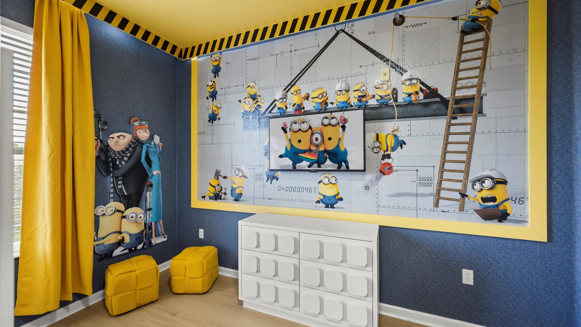 Minions-themed kids' bedroom featuring a large wall mural with various Minions characters, a wall-mounted TV, and a white dresser. Yellow curtains frame a window, and yellow poufs are on the light wood floor.