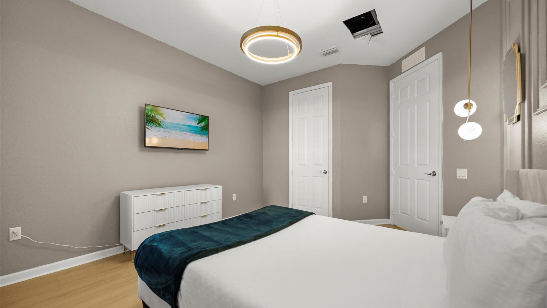 Modern bedroom with light beige walls, a white bed with a teal throw, a white dresser, and a wall-mounted TV displaying a beach scene. A circular light fixture hangs from the vaulted ceiling, illuminating the light wood floor.