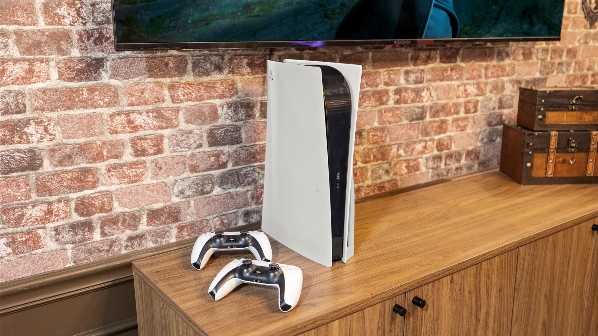 A close-up view in the Harry Potter-themed game room shows a PlayStation 5 console and two white controllers resting on a wooden media console. The console is set against a brick-patterned wall, beneath a large flat-screen TV displaying a movie scene.
