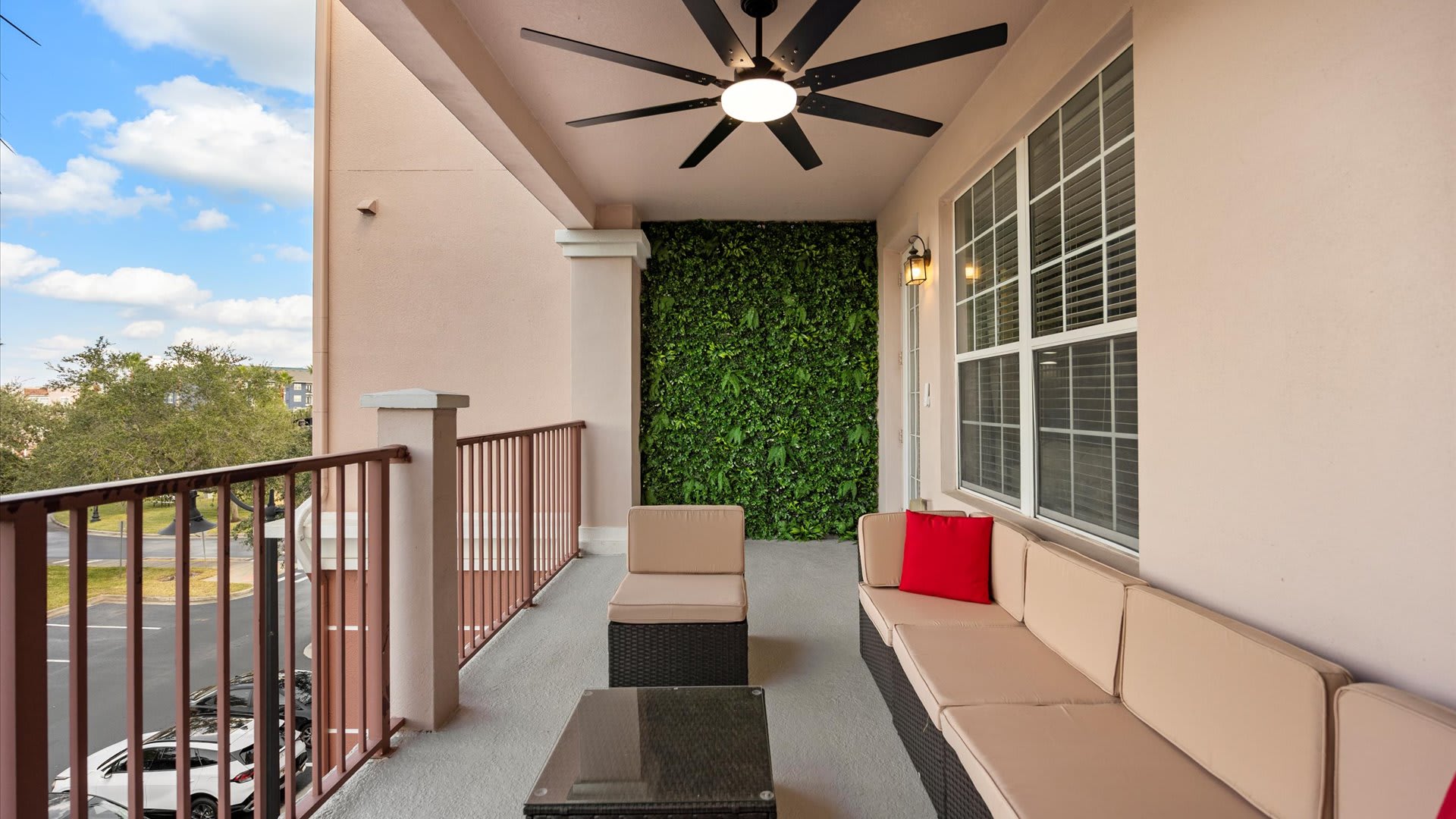 A wide balcony featuring outdoor wicker seating with beige cushions and a red accent pillow, under a black ceiling fan. A striking artificial green wall adds a natural touch, overlooking a parking lot and residential buildings.