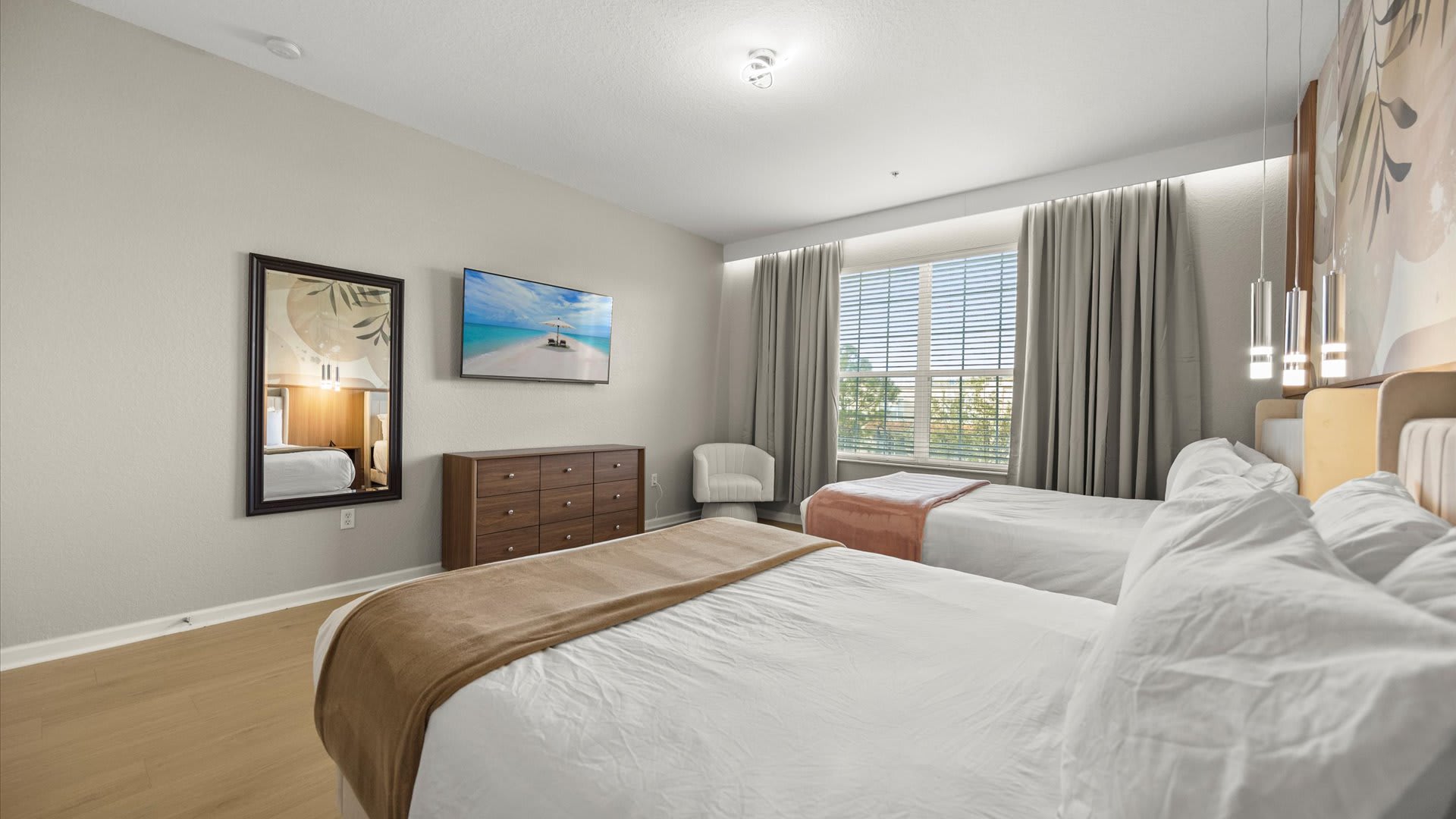 Wide shot of a bedroom with two queen beds, a large window with blinds and drapes, and a wall-mounted TV above a wooden dresser. A decorative accent wall with botanical patterns adds character to this bright room with light wood floors.