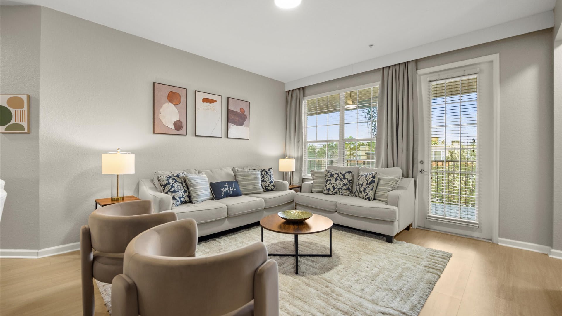 A bright living room furnished with two light-colored sofas and two beige armchairs, arranged around a round wooden coffee table on a textured area rug. The room is decorated with abstract artwork and features large windows and a glass door leading to a balcony.