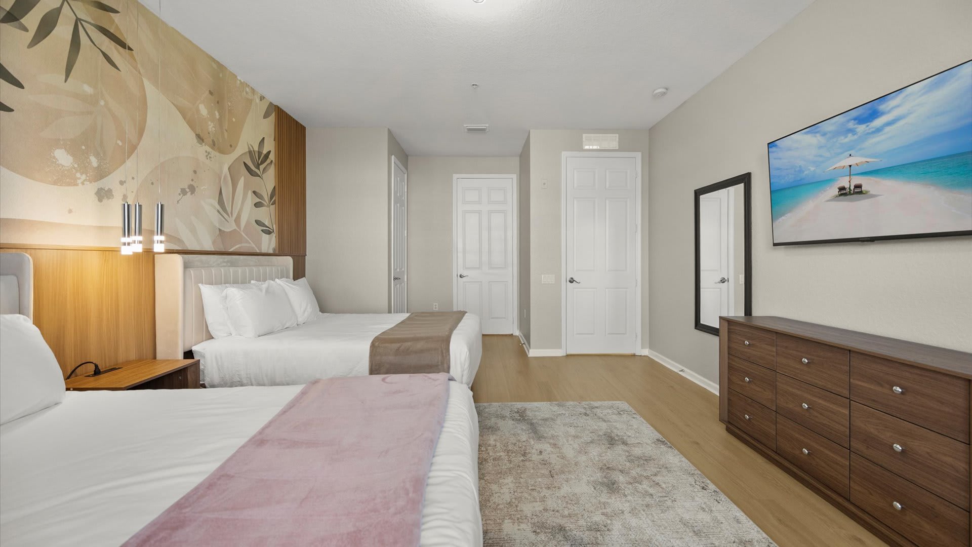 View of a spacious bedroom with two queen beds, a wooden dresser with a wall-mounted TV, and a full-length mirror. The accent wall behind the beds features a botanical design, adding a touch of nature to the room with light wood floors.