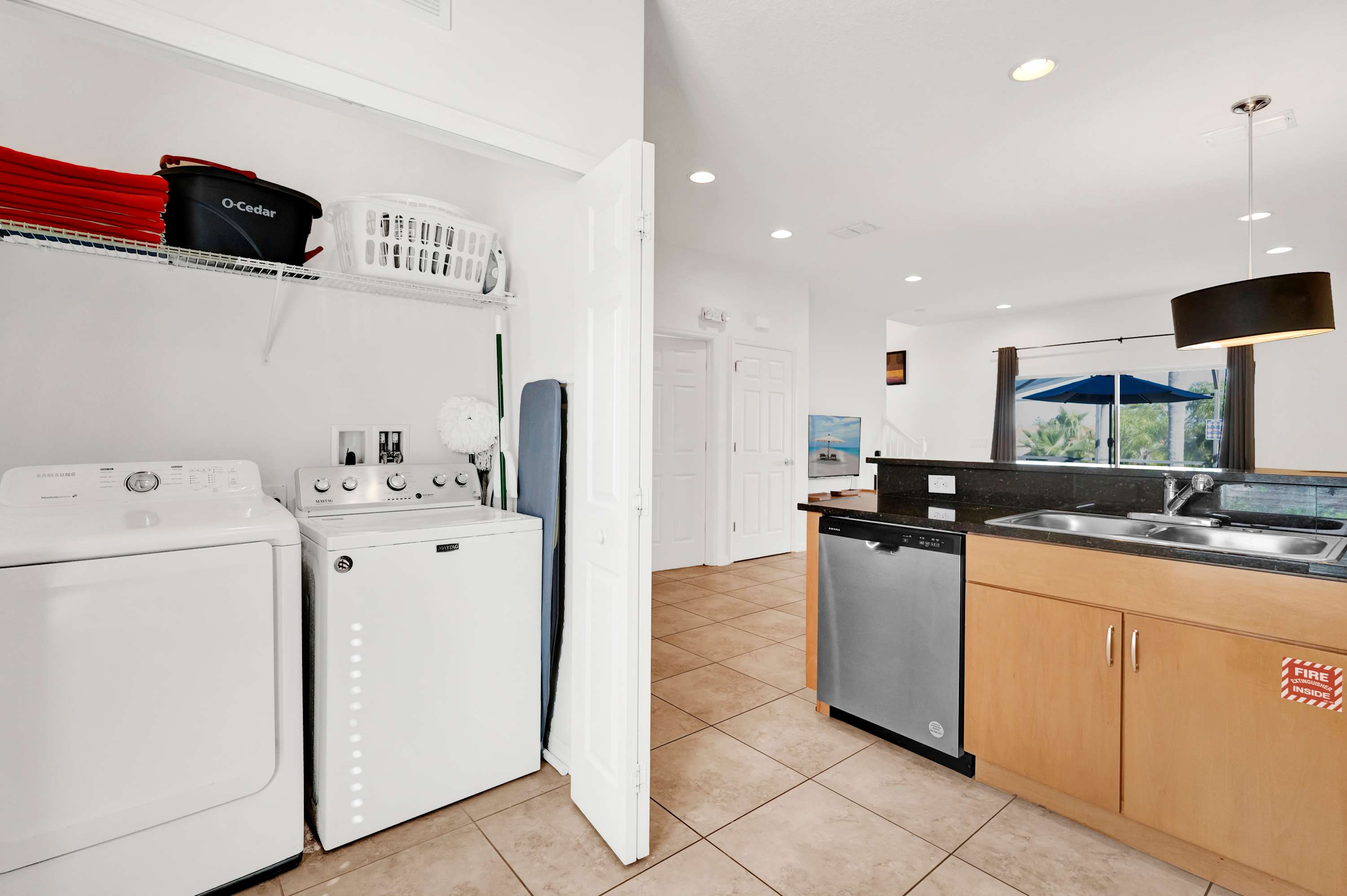 A dedicated laundry area features a white top-loading washing machine and a front-loading dryer, with shelving above holding laundry baskets and supplies. Adjacent is a kitchen counter with a sink and dishwasher.