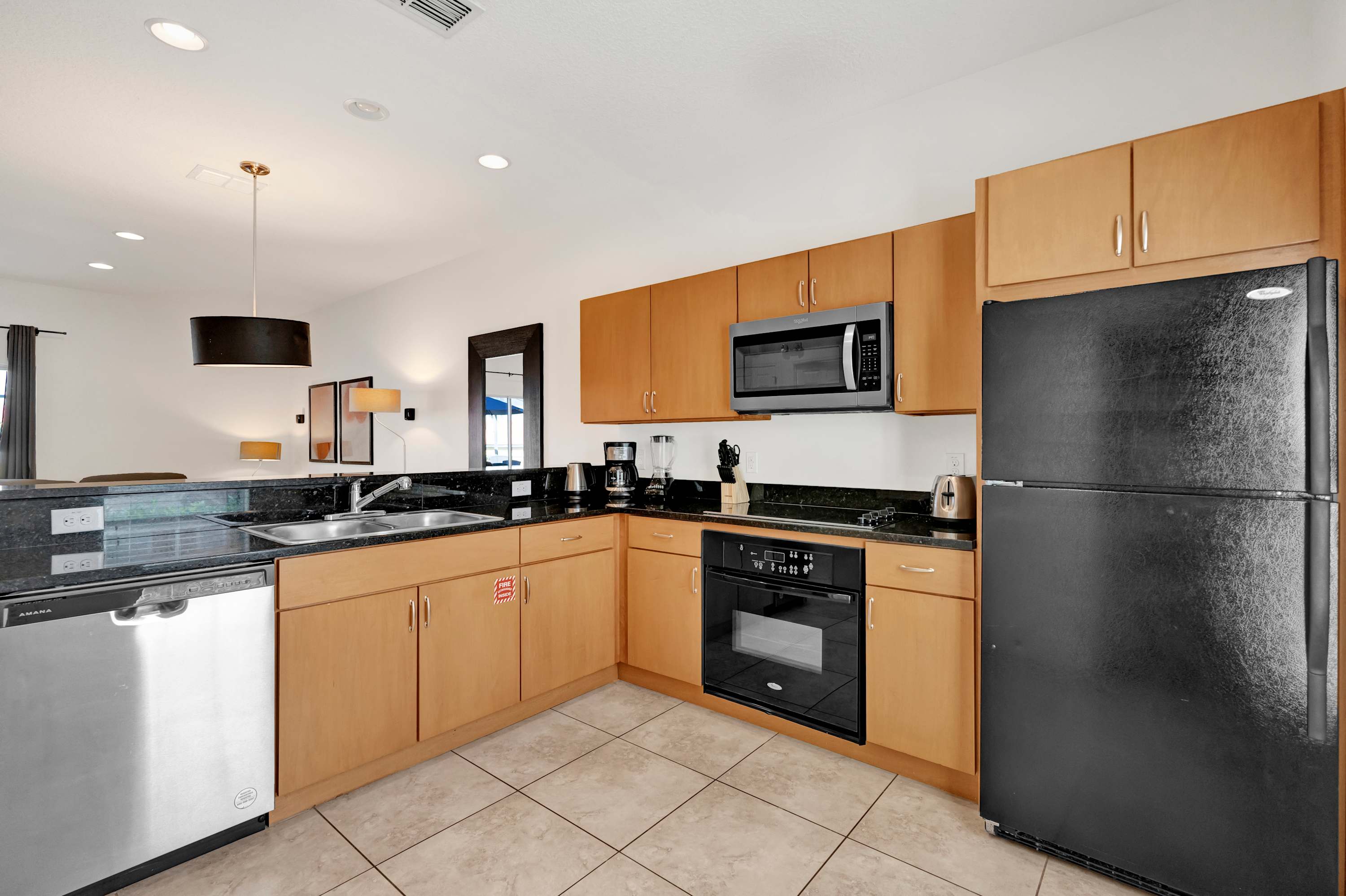 Wide shot of the modern kitchen featuring light wood cabinets, black granite countertops, and stainless steel appliances including a microwave, oven, and a black refrigerator. A stainless steel dishwasher is visible next to the double basin sink.