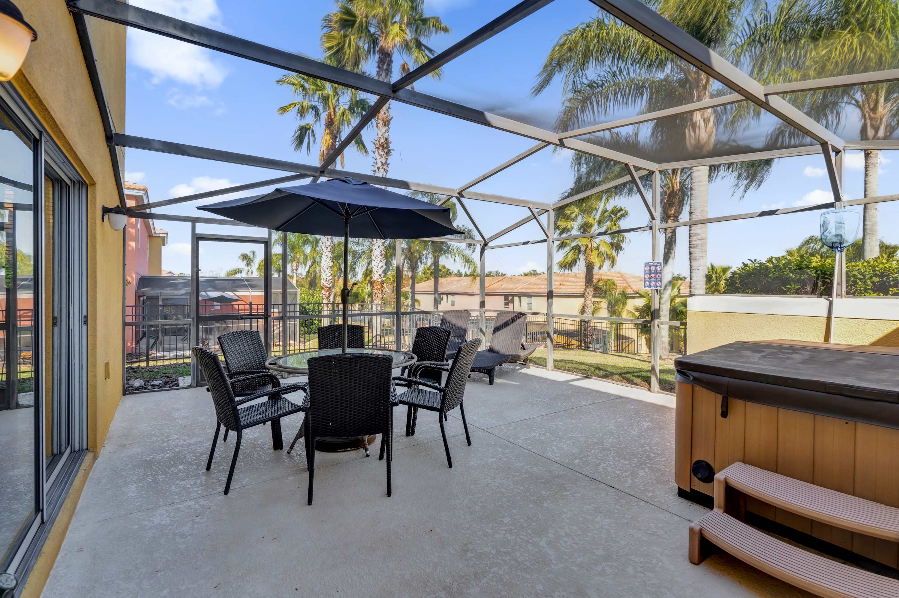 A spacious screened outdoor patio features a hot tub with wooden steps, a glass-top dining table with four black wicker chairs, and two black lounge chairs. The light concrete floor extends under the metal screen enclosure, with palm trees and distant houses visible.