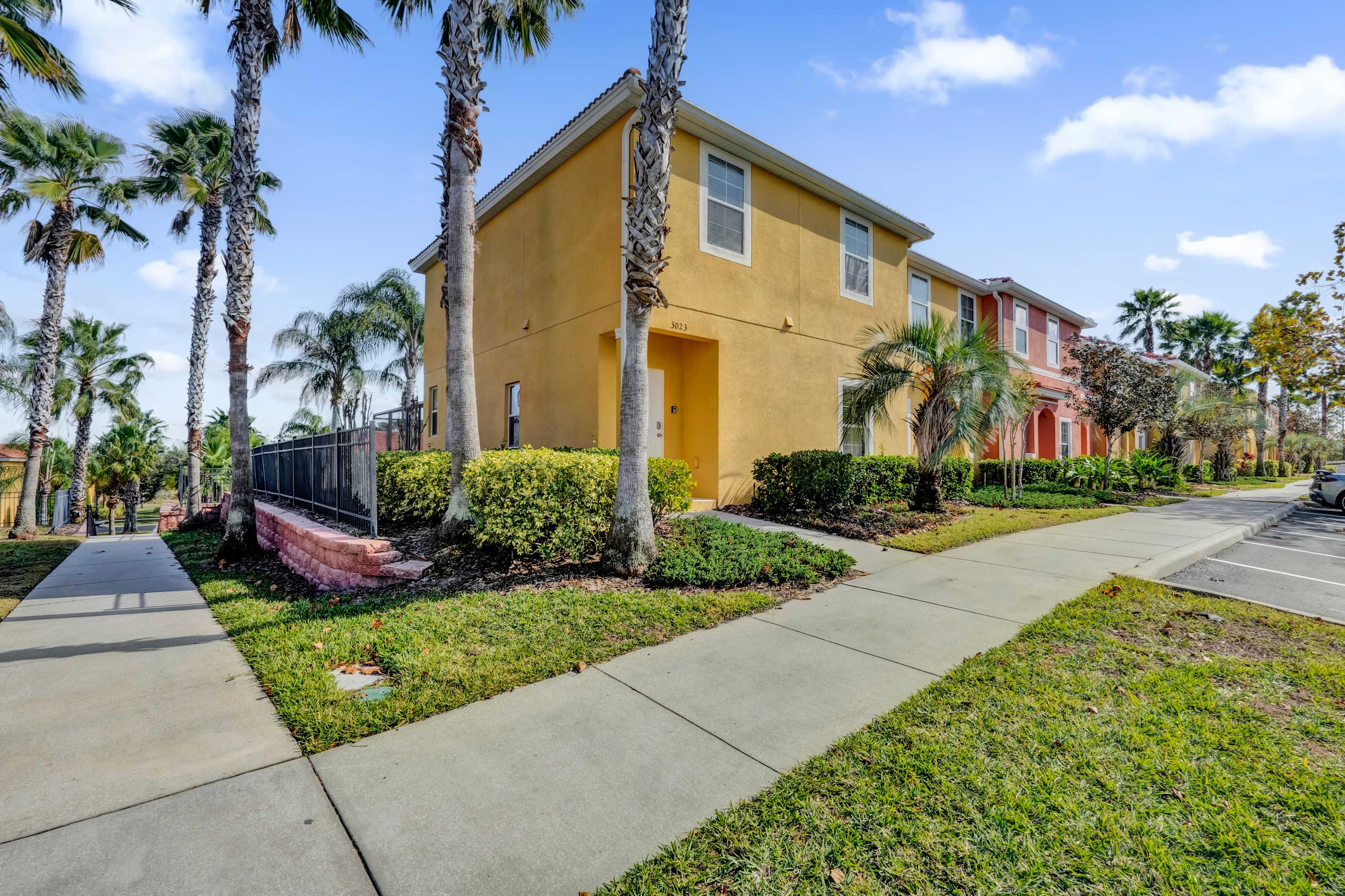 The exterior of a two-story yellow townhouse features a white front door and multiple windows, surrounded by green grass, hedges, and tall palm trees. A concrete sidewalk leads to the entrance, with other colorful townhouses visible in the background.