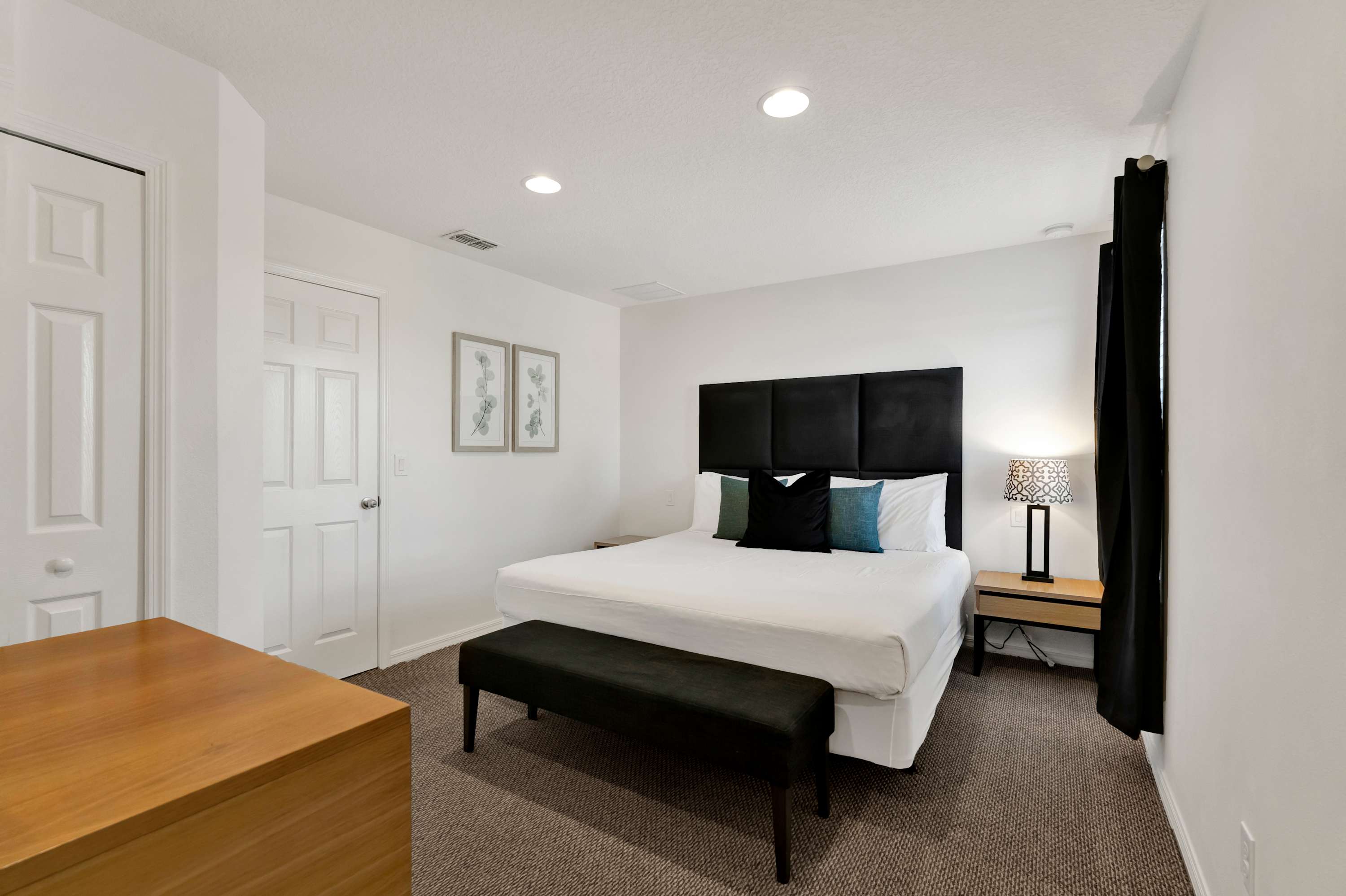 Spacious bedroom with a king-sized bed featuring a black upholstered headboard, white bedding, and accent pillows. A dark wood nightstand with a lamp is on the right, and a black bench sits at the foot of the bed.