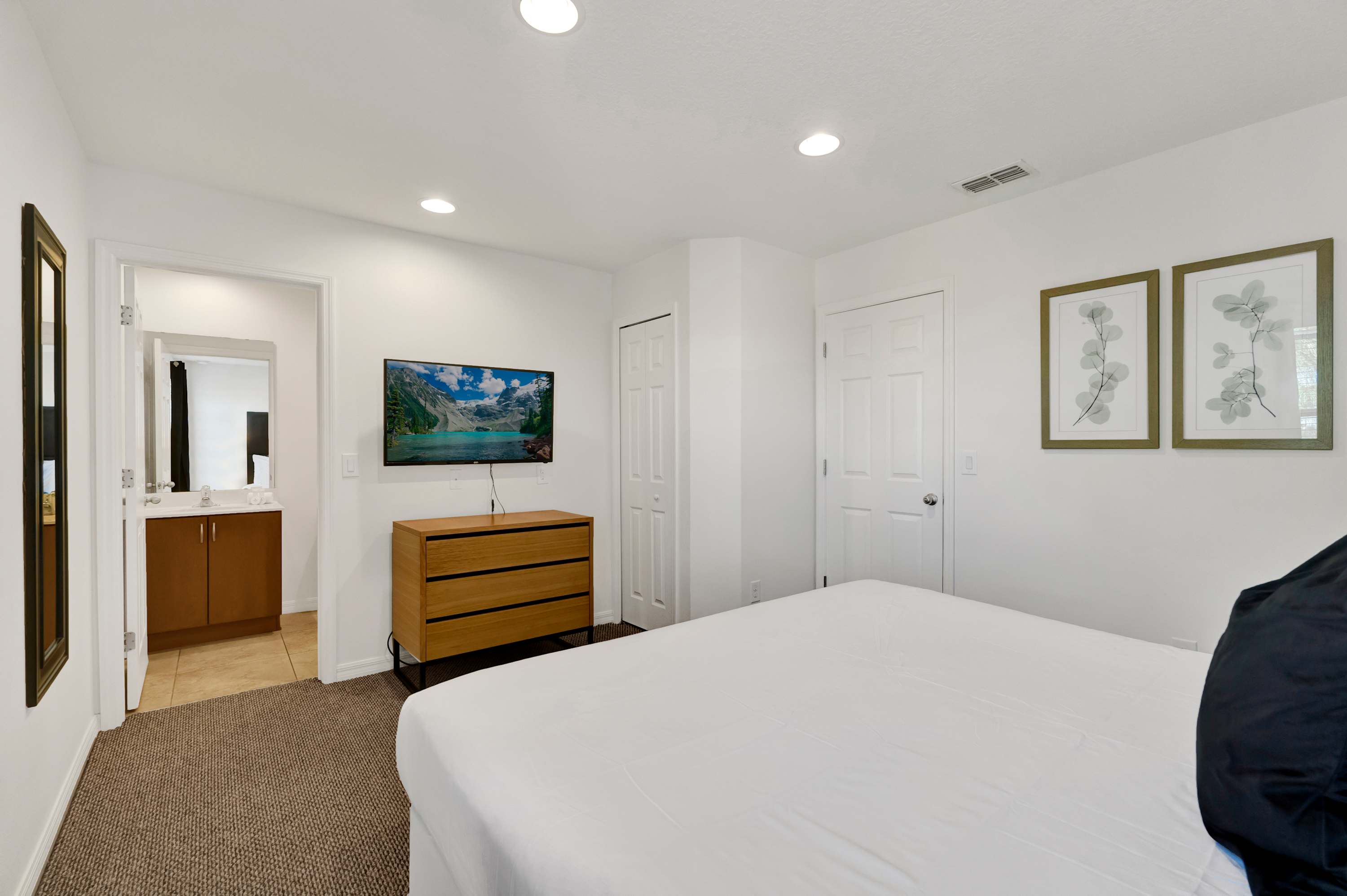 Another view of the first bedroom, showing a wall-mounted TV above a wooden dresser. A doorway leads to an en-suite bathroom, and two framed botanical prints adorn the wall.