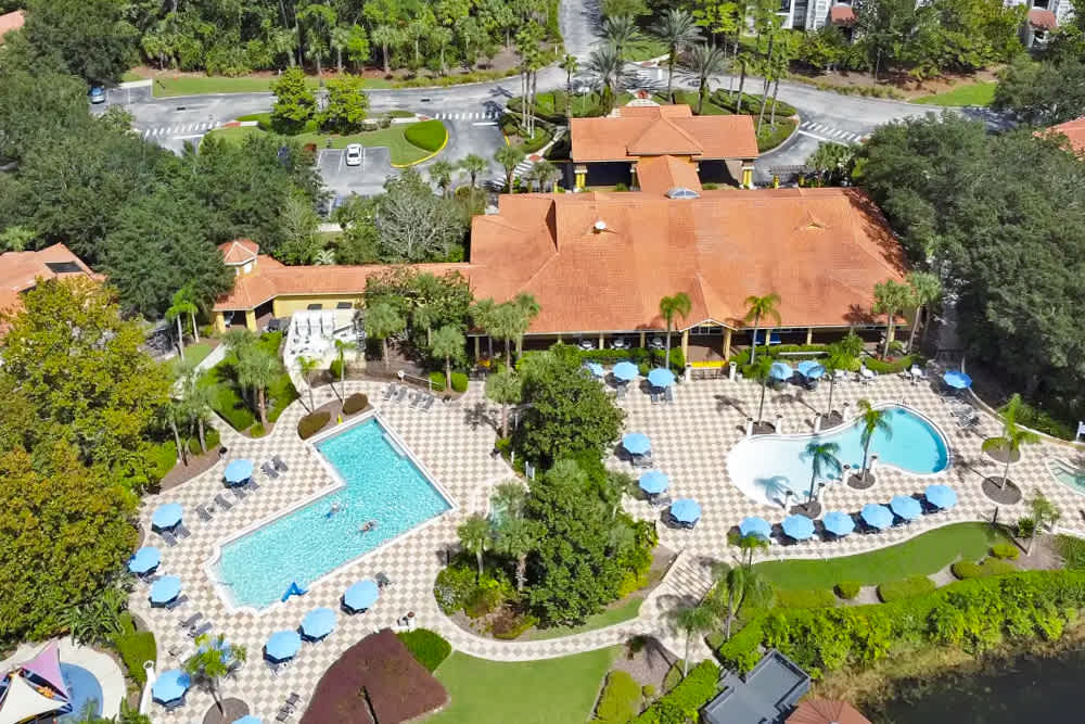 An aerial view showcases a large resort complex with multiple swimming pools, surrounded by numerous blue umbrellas and lounge chairs on a paved deck. Lush green landscaping and palm trees separate the pools from a large building with a terracotta roof and surrounding parking areas.