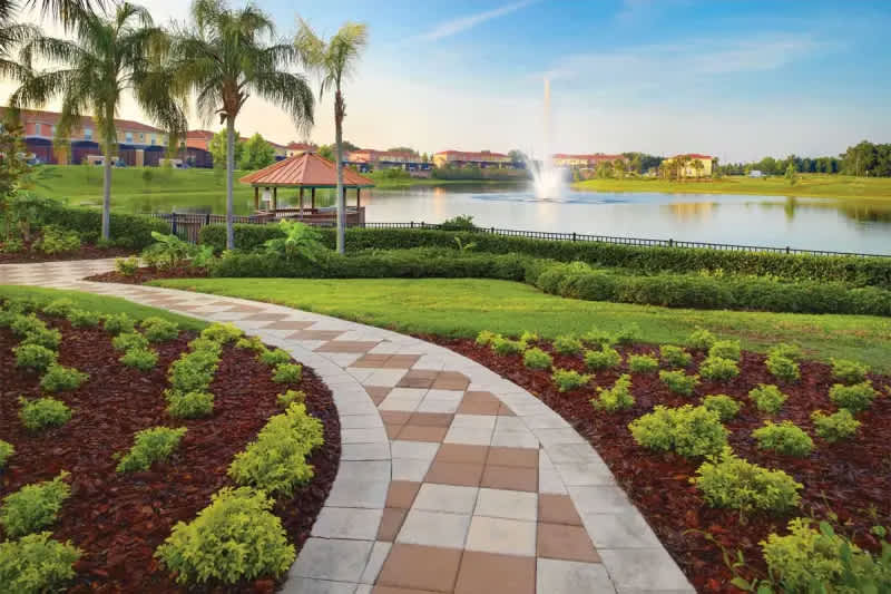 A picturesque outdoor garden area featuring a winding paved path with a checkered pattern, surrounded by lush green grass and landscaped beds with small shrubs and mulch. In the background, a serene lake with a fountain and a charming gazebo are visible.