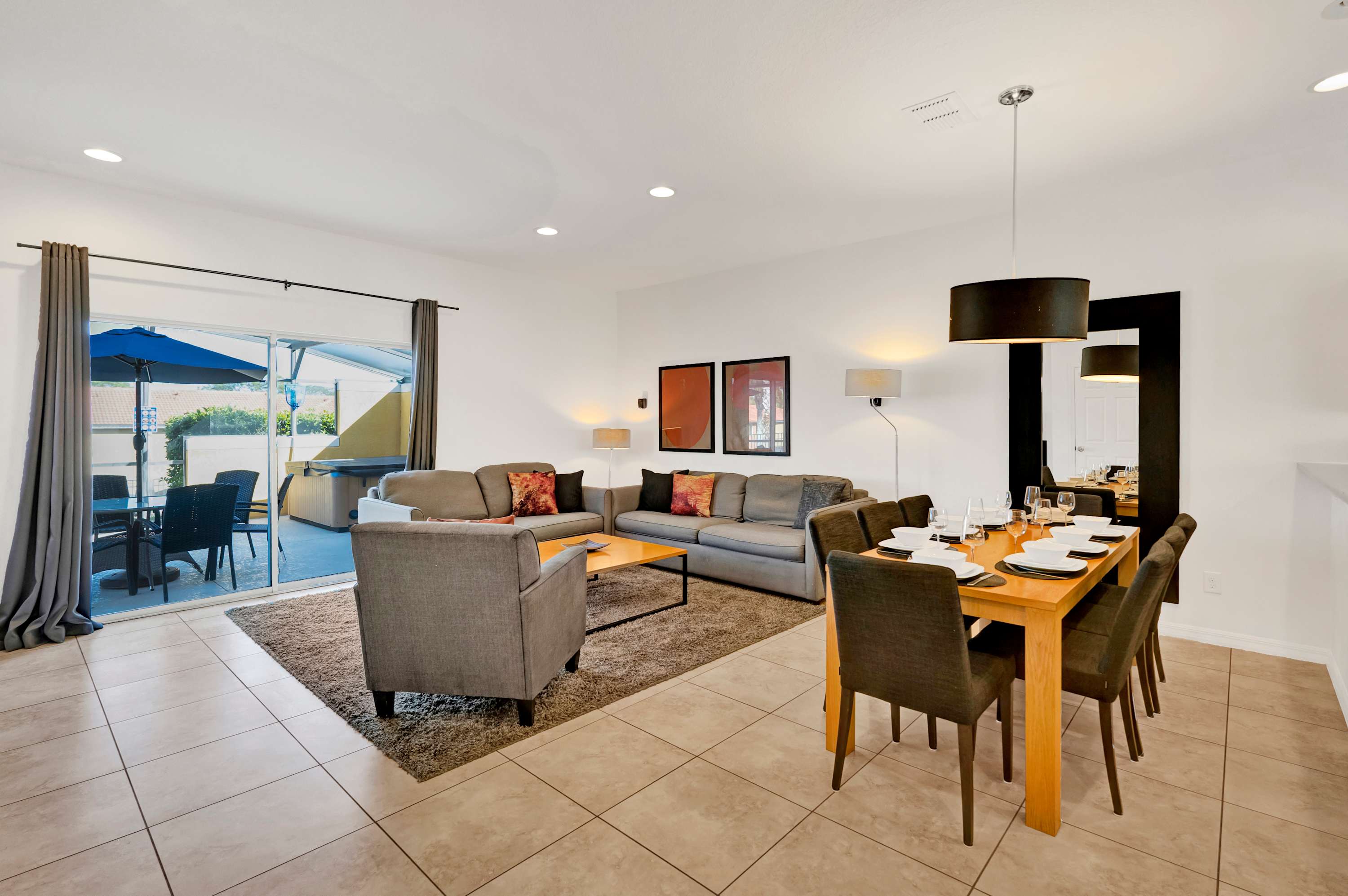 An open-concept living and dining area features a gray sectional sofa and armchair on a brown rug, adjacent to a light wood dining table set for six with dark upholstered chairs. Sliding glass doors offer a view of the outdoor patio, and the space is illuminated by recessed lighting and a modern pendant lamp.