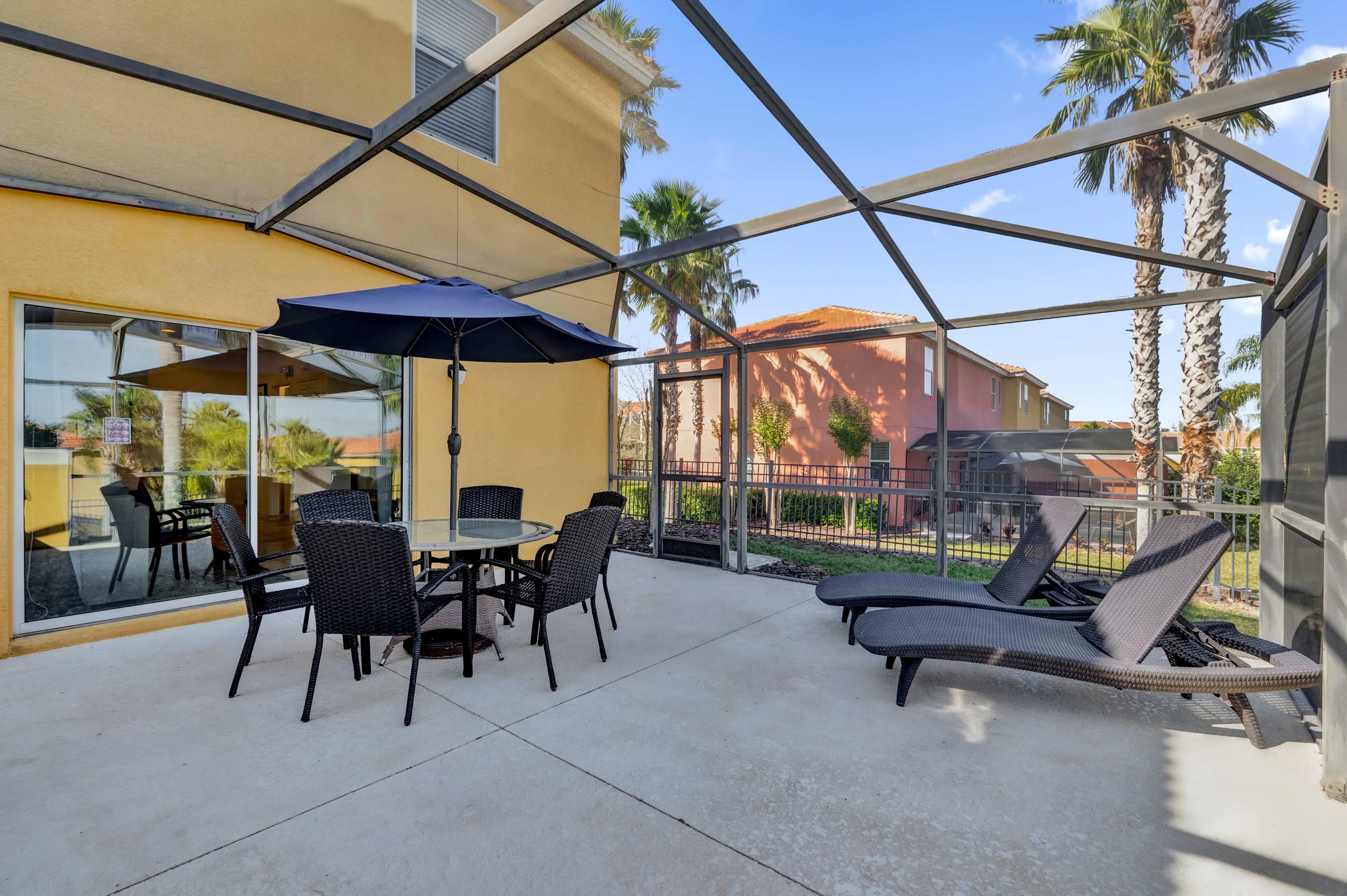 A screened outdoor patio features a glass-top dining table with four black wicker chairs and two black lounge chairs on a light concrete floor. The screen enclosure offers views of palm trees and other residential buildings in the distance.
