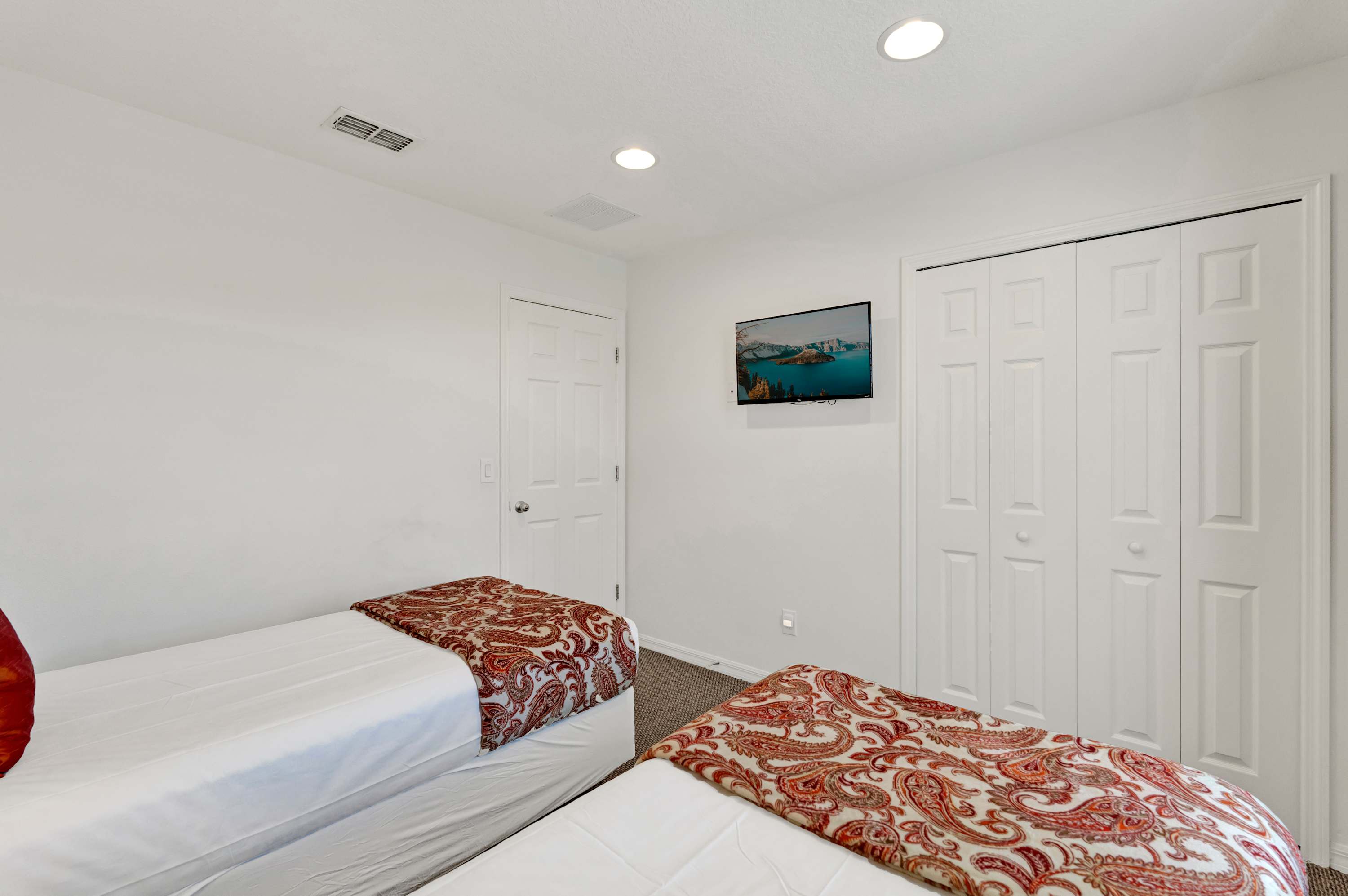 Another view of the third bedroom, showing the two twin beds, a wall-mounted TV, and bi-fold closet doors. The room has white walls and a carpeted floor.