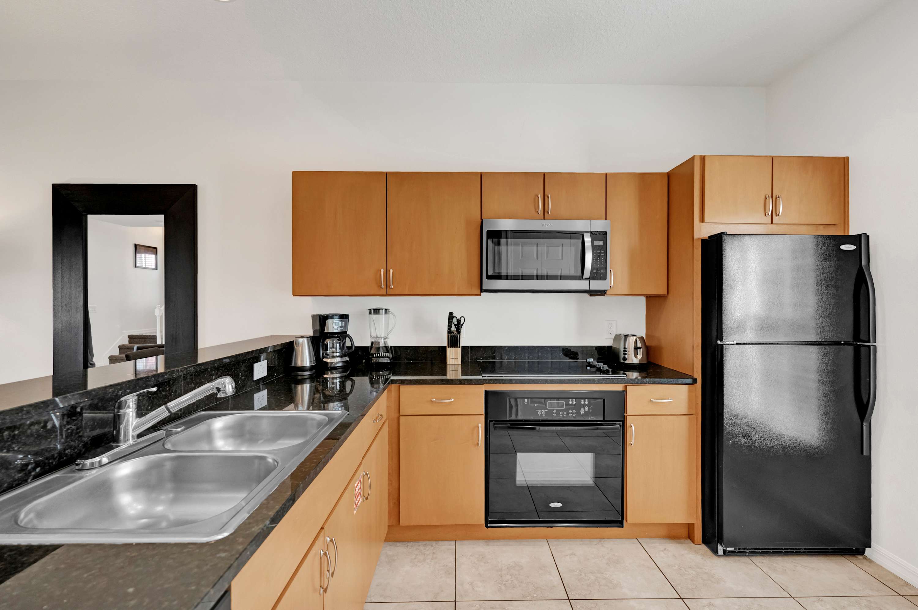 Modern kitchen with light wood cabinetry, black granite countertops, and stainless steel appliances including a microwave, oven, and a black refrigerator. A double basin sink is visible on the left.
