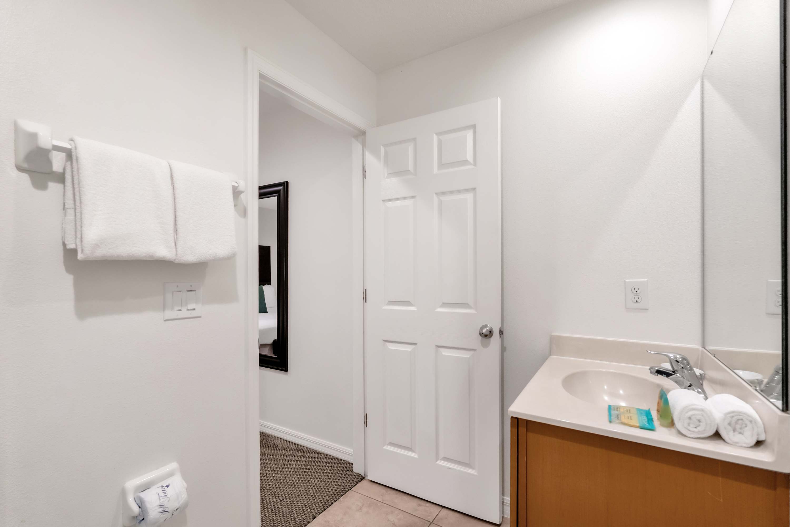 A bright bathroom features a light-colored vanity with a sink, a large mirror, and a towel rack holding white towels. An open door reveals a glimpse of an adjacent room with a dark mirror.