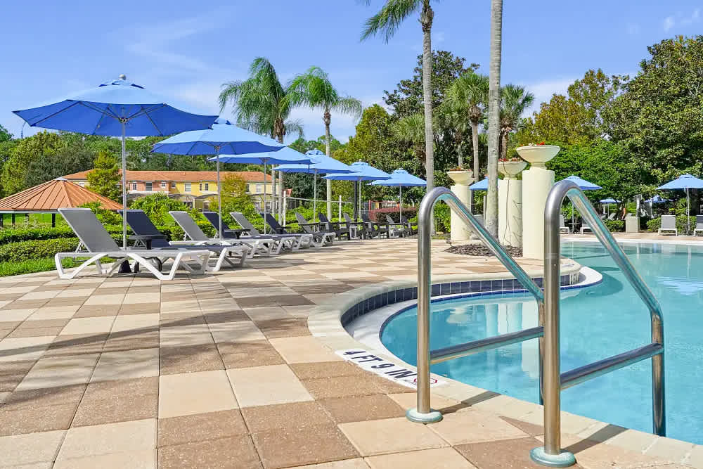 A spacious communal swimming pool area features numerous lounge chairs with blue umbrellas on a checkered paved deck. Palm trees and lush greenery surround the inviting pool.