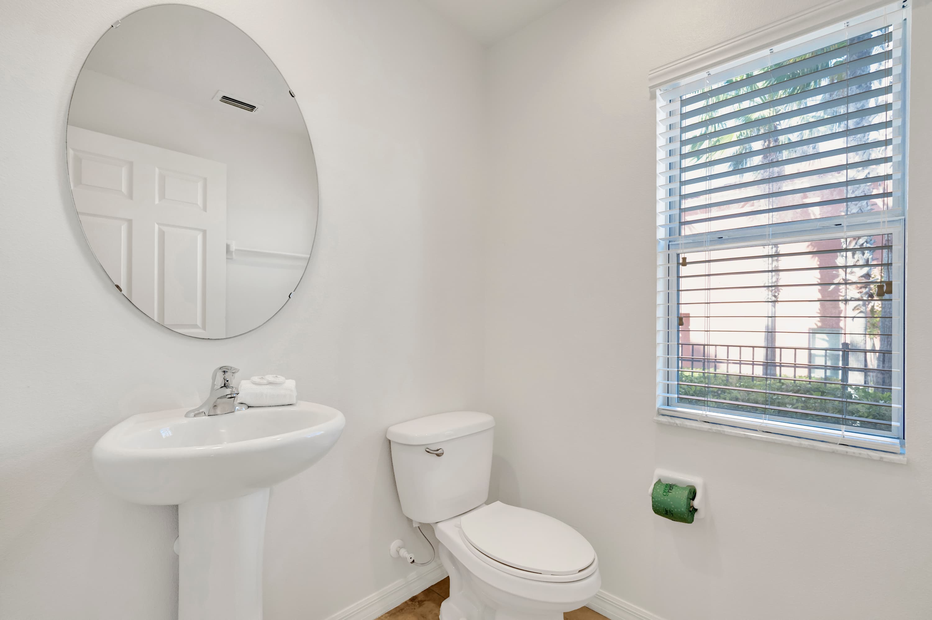 A bright half-bathroom features a white pedestal sink, a toilet, and a large round mirror above the sink. A window with white blinds allows natural light, and a green toilet paper holder is mounted on the wall.