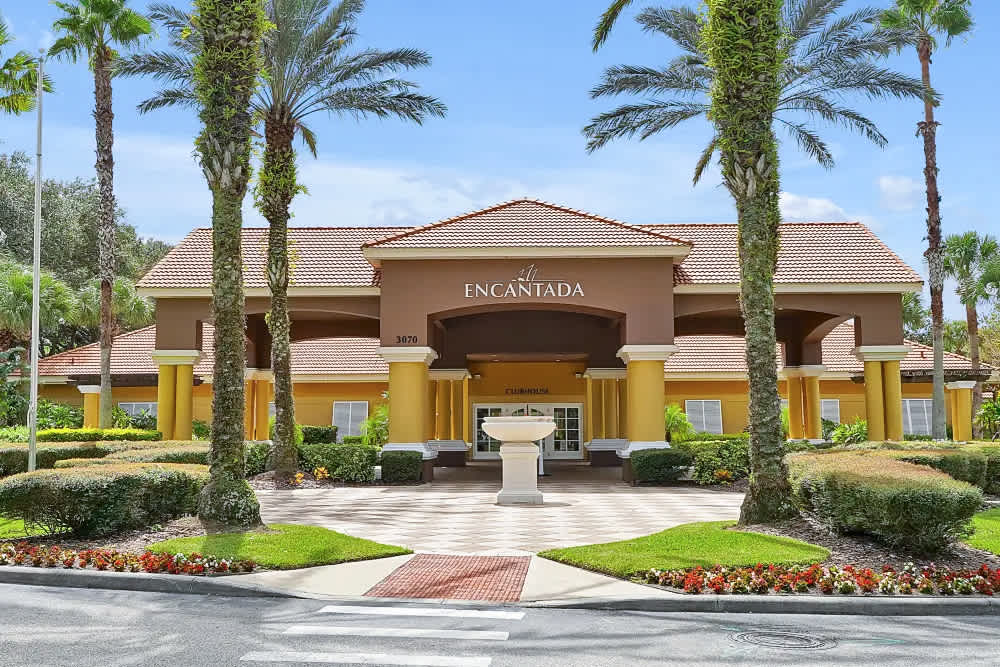 The grand entrance of "La Encantada" clubhouse features a Spanish-style building with a red tile roof, yellow columns, and lush landscaping. A fountain stands in the paved courtyard.