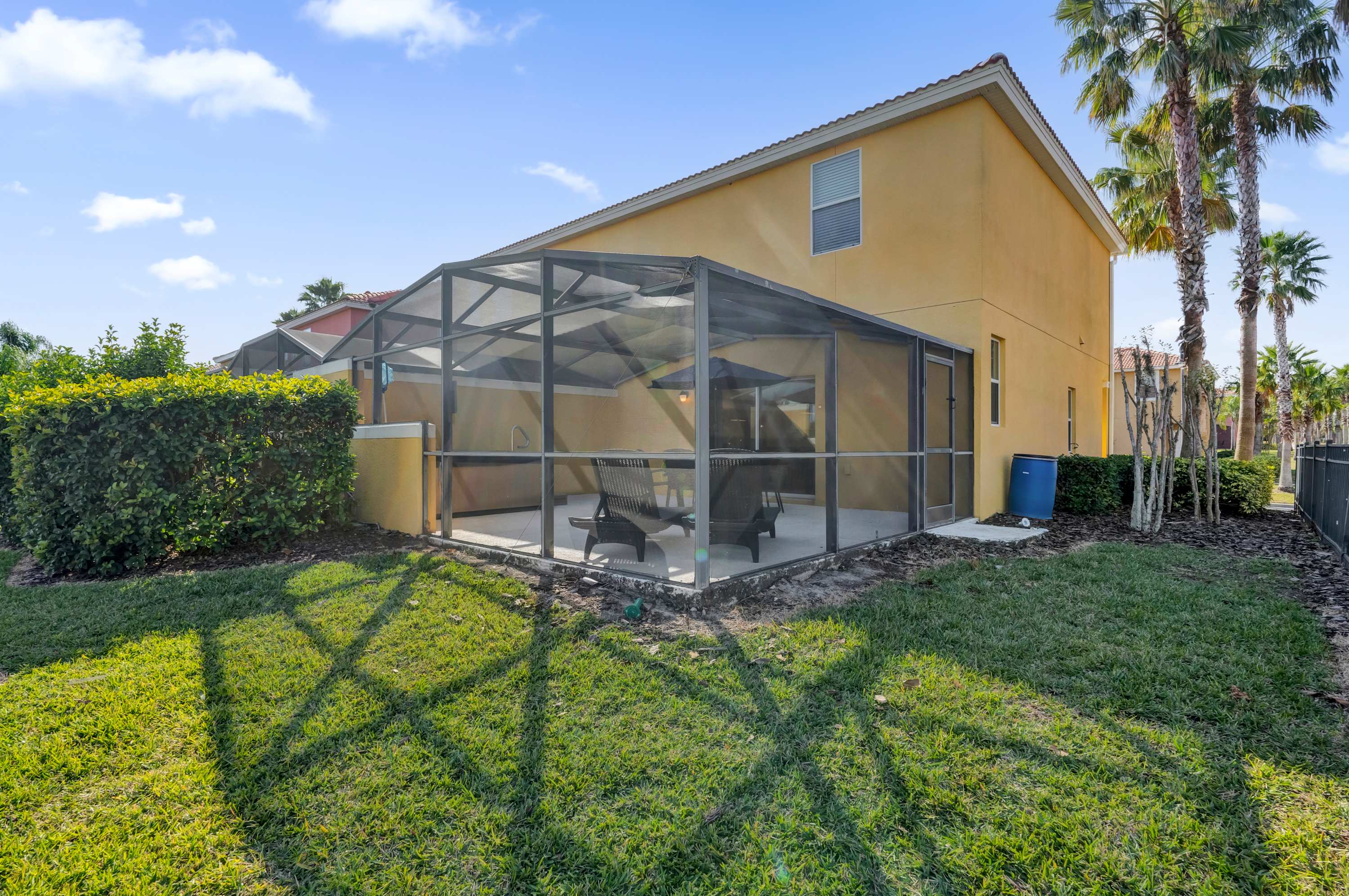 The exterior back of the townhouse features a screened-in patio with outdoor seating, overlooking a well-maintained green lawn. The yellow-painted house has a second-story window and is surrounded by lush landscaping.