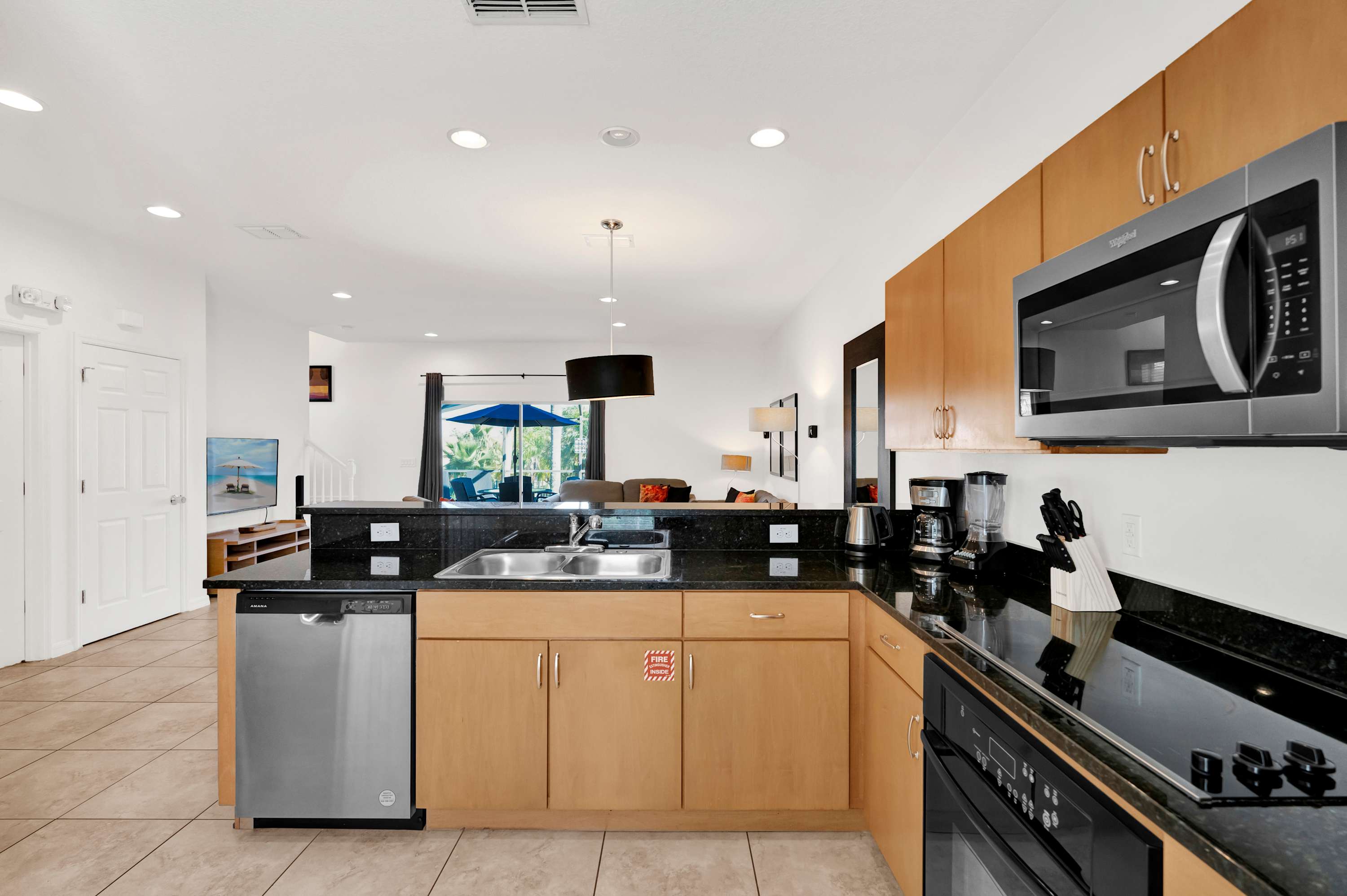 View of the kitchen from a different angle, showing the double basin sink, stainless steel dishwasher, and a partial view of the living area beyond the counter. The kitchen features light wood cabinets and black granite countertops.