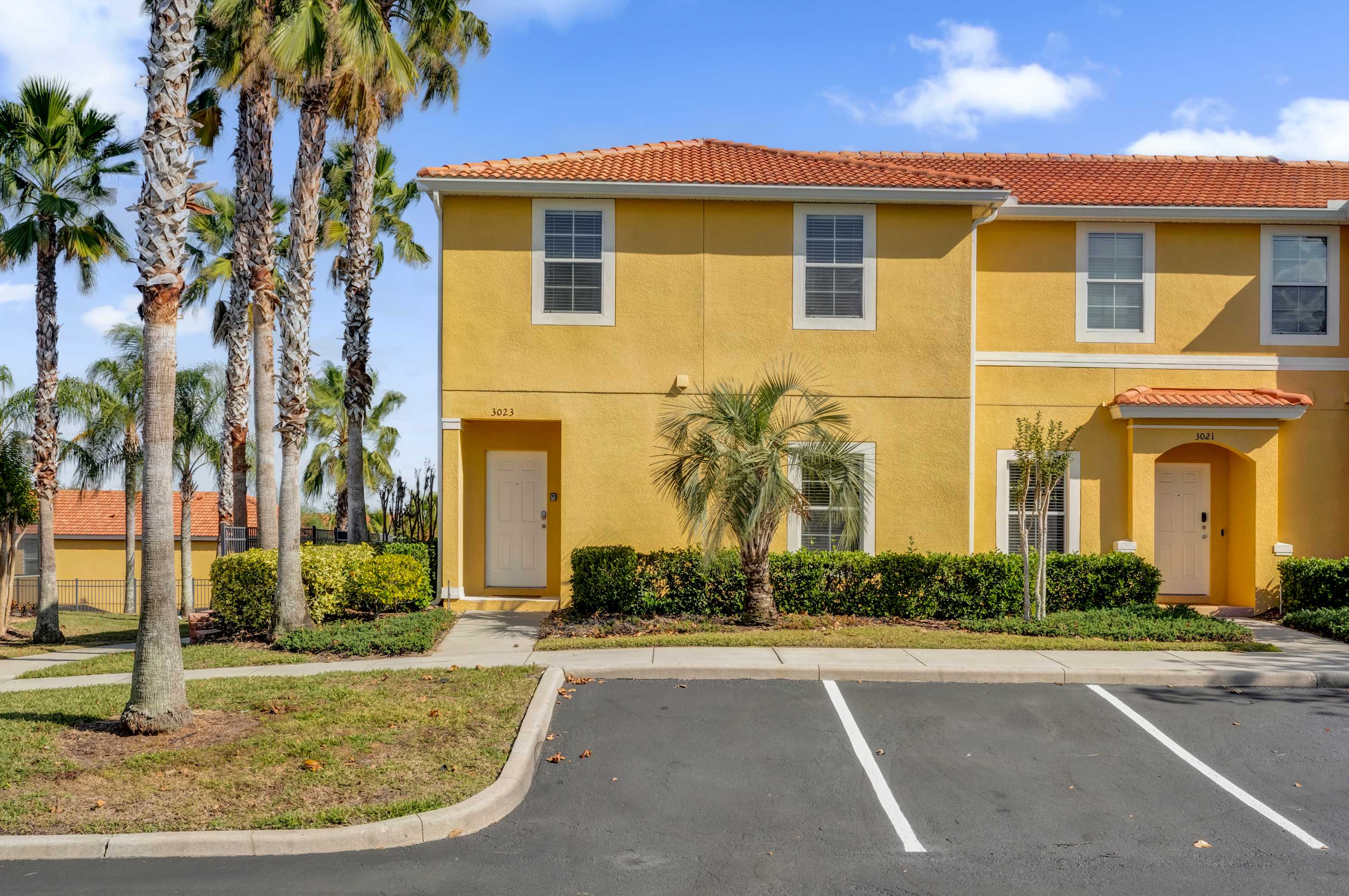 The front exterior of a yellow townhouse with a red tile roof features a white front door and multiple windows with white blinds. Palm trees line the property, and paved parking spaces are visible in the foreground.