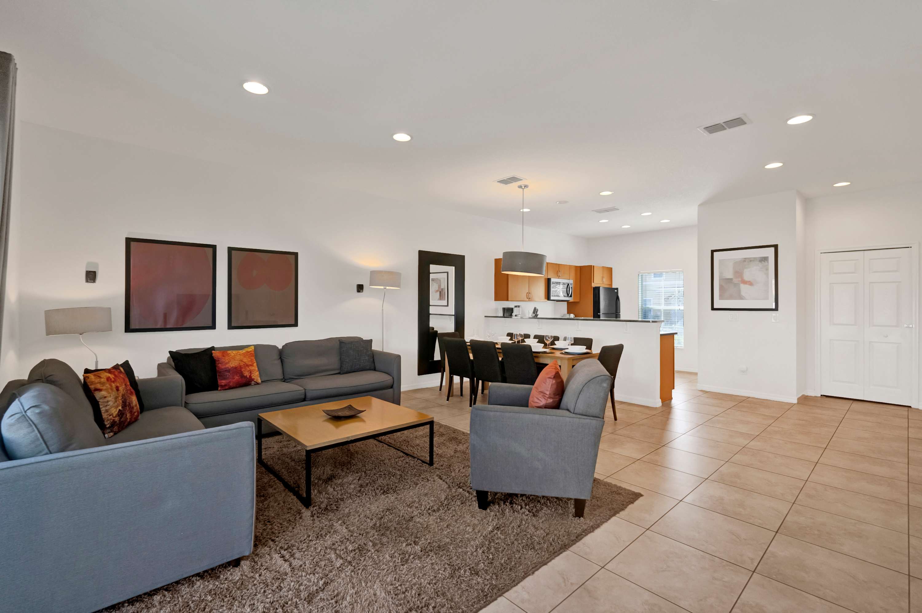 A bright, open-concept living and dining area features a large gray sectional sofa and armchair on a shaggy brown rug, facing a wooden coffee table. A light wood dining table with dark chairs is visible to the right, and sliding glass doors lead to an outdoor patio.