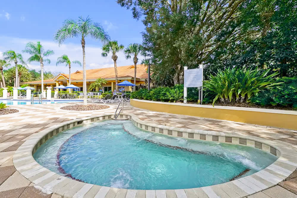 A bubbling hot tub with tiled edges is set within a paved deck, surrounded by lush tropical landscaping and palm trees. The clubhouse building is visible in the background.