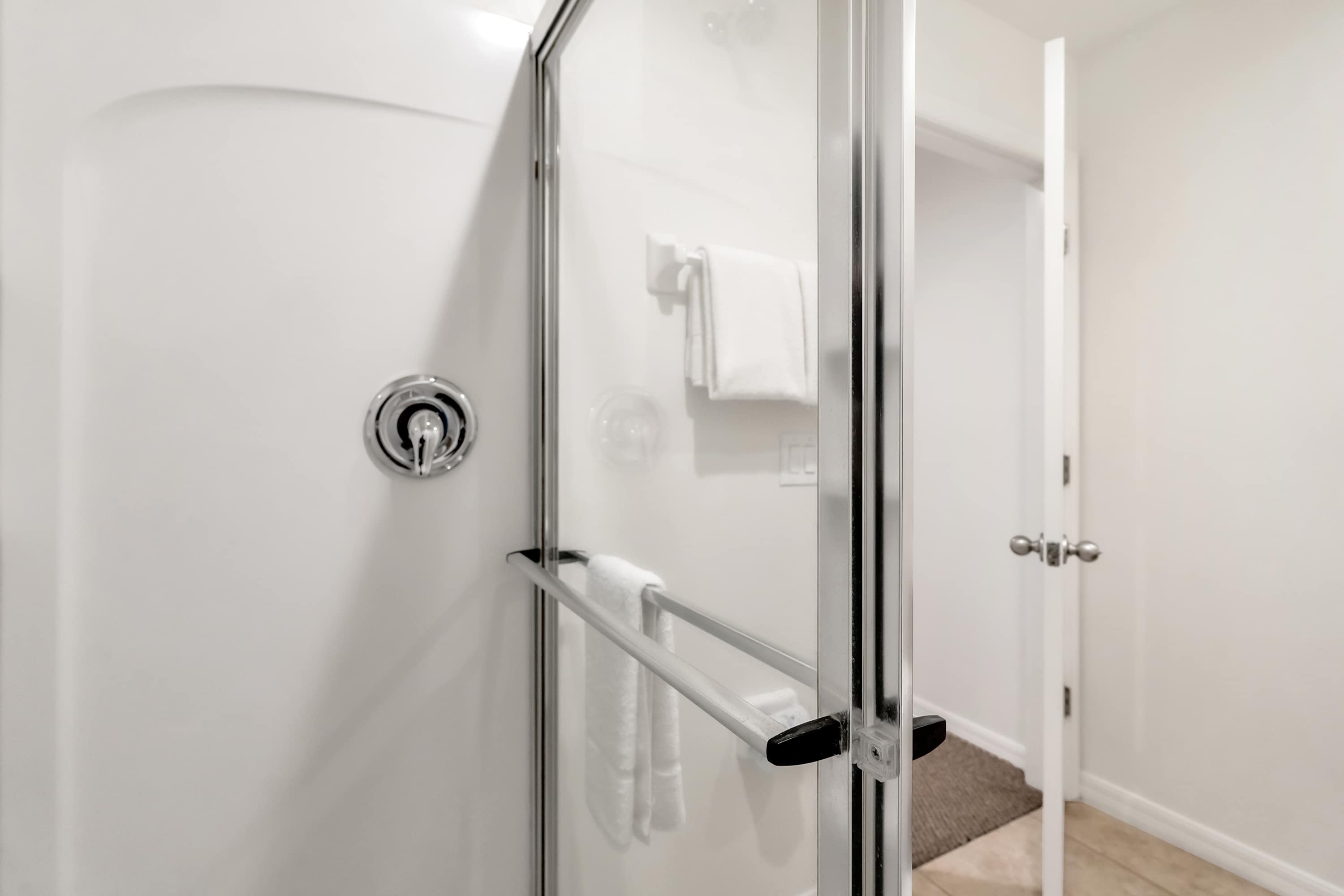 A modern shower stall with clear glass doors and a chrome shower head is visible, featuring white tiled walls and a grab bar. White towels are hanging on a rack inside the shower.