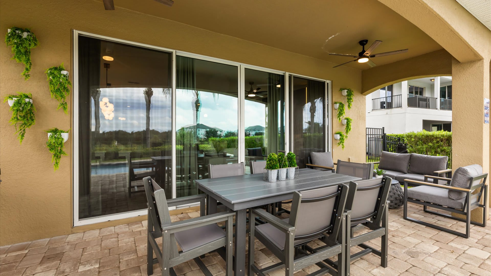 A spacious covered patio features a modern outdoor dining set and a lounge area with comfortable seating, all situated on a paver patio. Large sliding glass doors connect to the interior of the house.