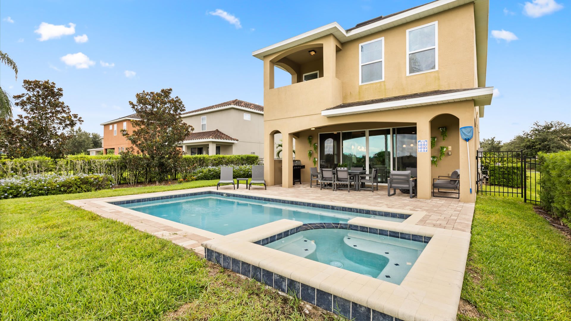 The back exterior of a two-story house with a private rectangular swimming pool and an integrated hot tub, surrounded by a green lawn and a low hedge. A covered patio with outdoor furniture is visible next to the house.