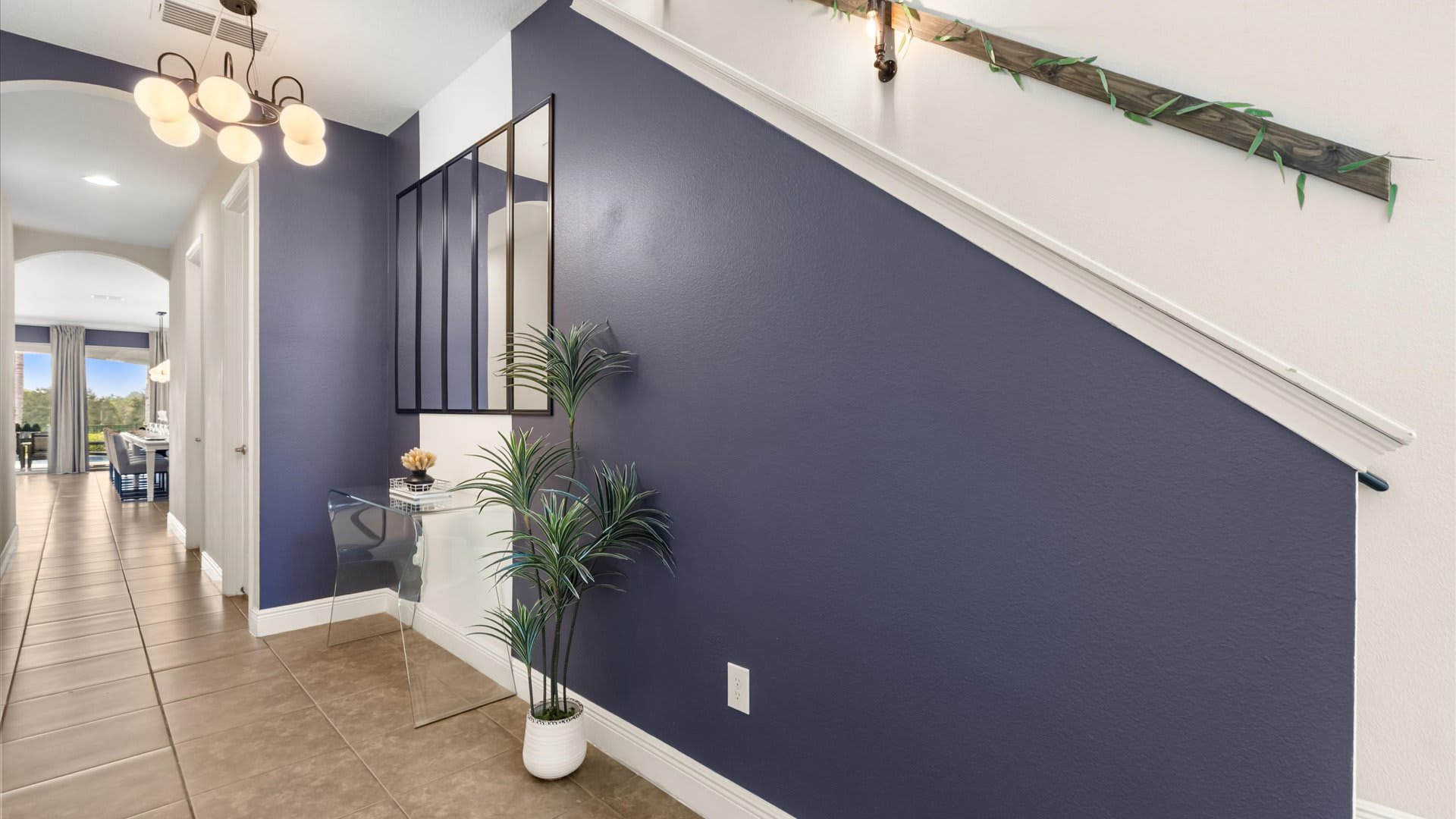 A vibrant entryway with deep purple walls and a light-colored staircase featuring a decorative wooden railing. The space includes a modern console table with a plant, a large mirror, and a multi-bulb chandelier.