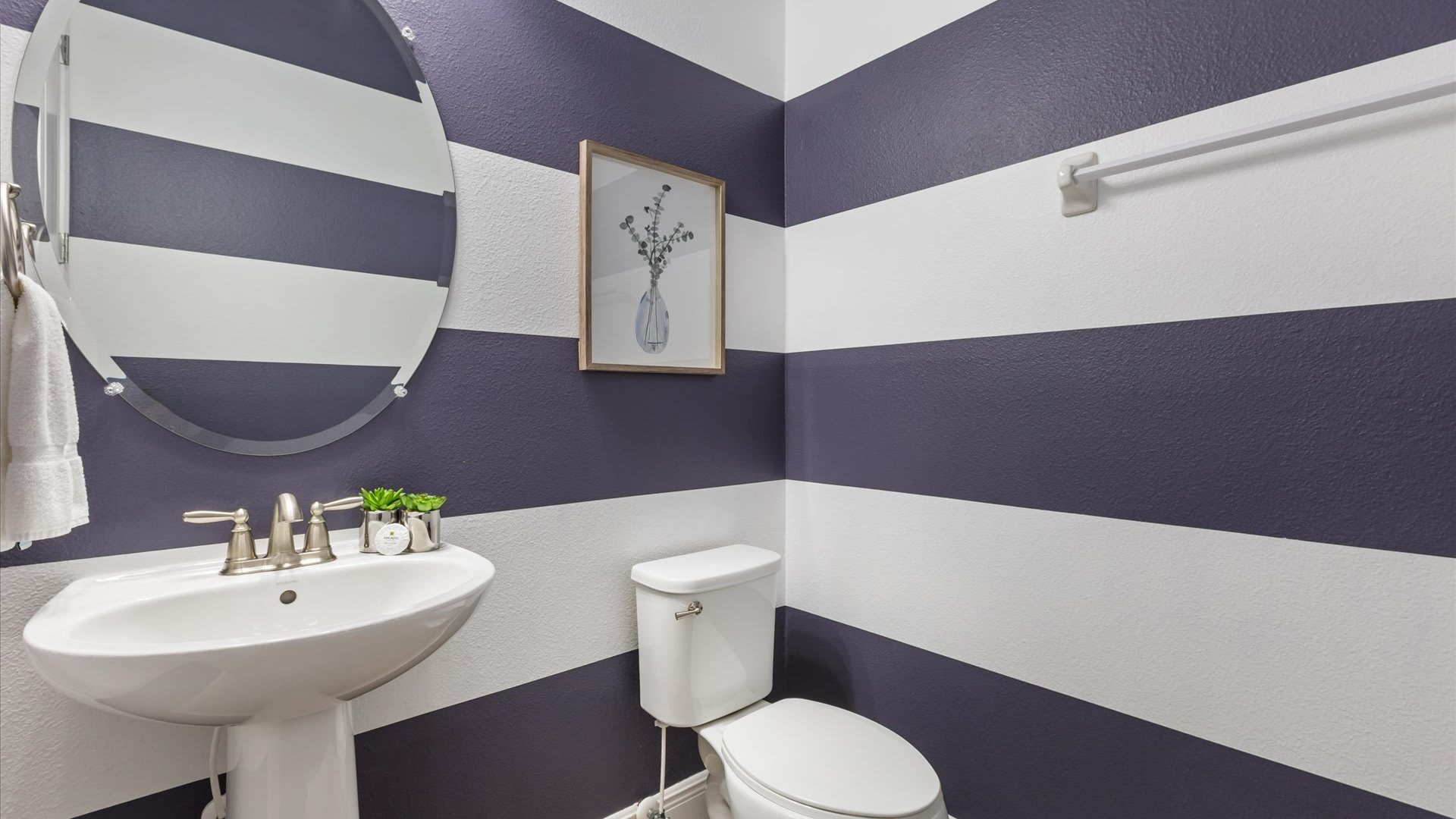 A stylish powder room with bold horizontal purple and white striped walls, featuring a white pedestal sink with a brushed nickel faucet and an oval mirror. A white toilet and a small framed botanical print complete the space.