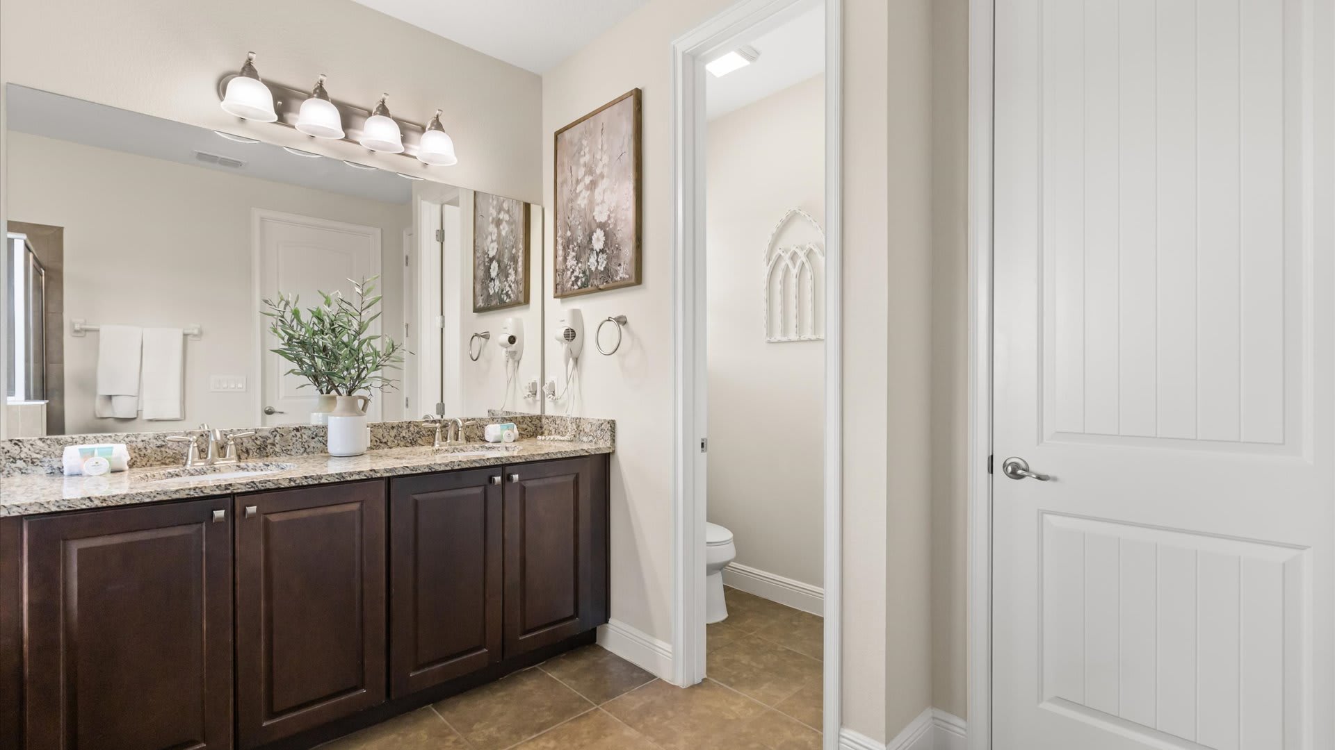 A bright bathroom with a double vanity featuring granite countertops and dark wood cabinets, a large mirror, and a separate alcove for the toilet.