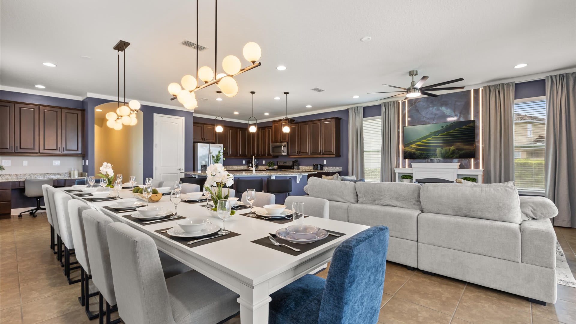 An expansive open-concept space features a long dining table set for a large group under modern globe chandeliers, with a view of the kitchen island and a living area with a TV above a fireplace.