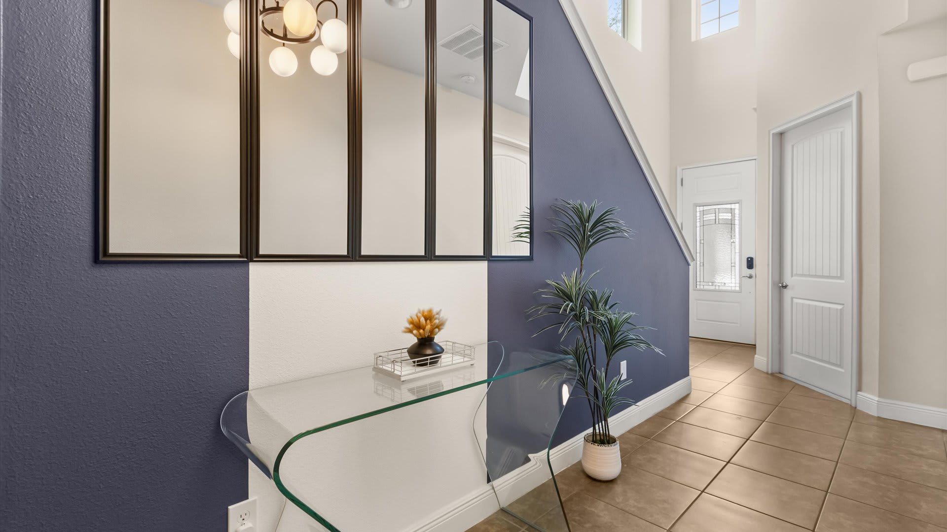 Bright foyer with a modern glass console table, decorative plant, and a series of black-framed mirrors on a two-toned wall. The space features light tiled flooring and a staircase leading upstairs.
