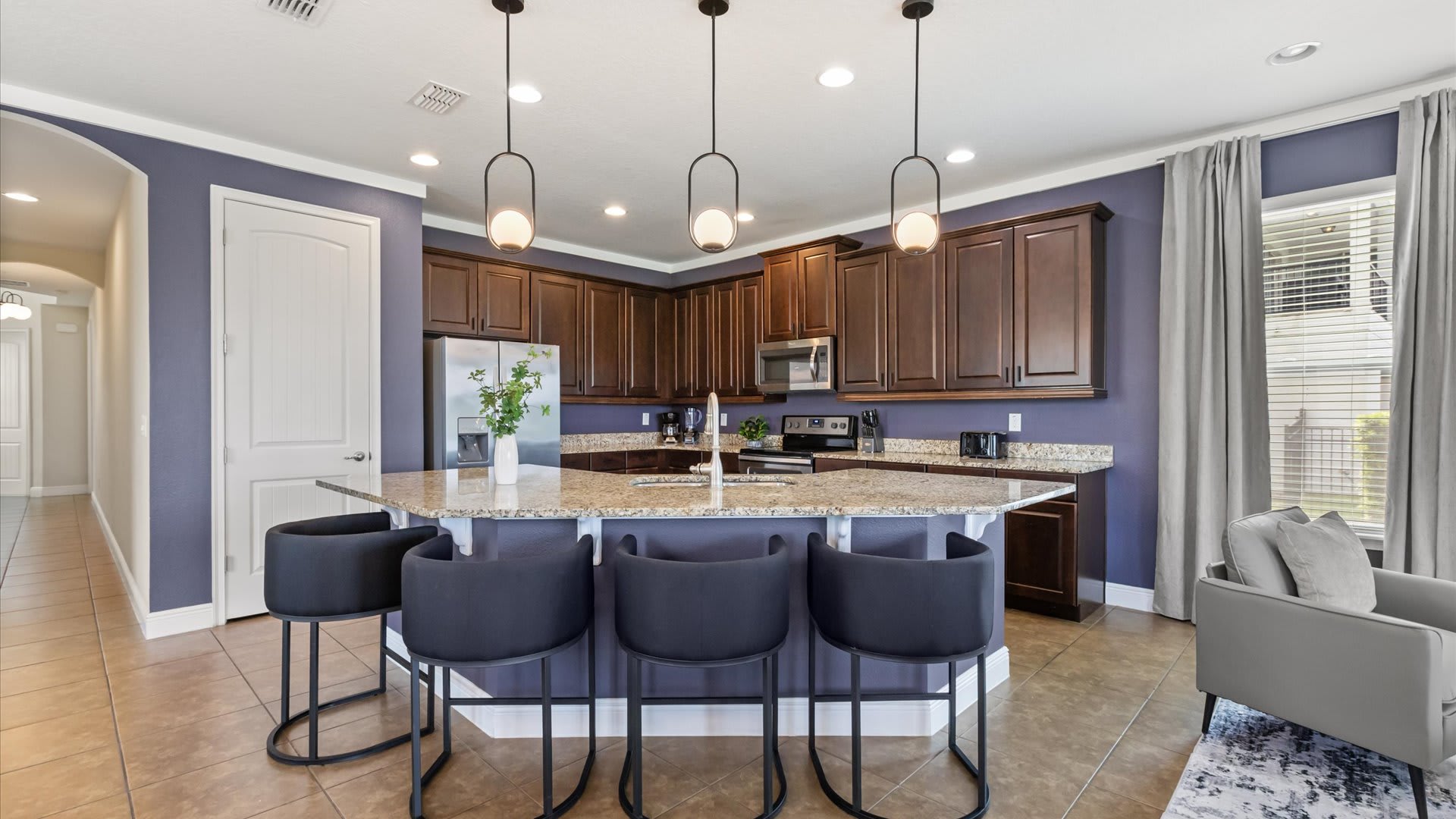 A well-appointed kitchen features a large island with three black bar stools, dark wood cabinetry, granite countertops, and stainless steel appliances including a refrigerator, microwave, and stove.