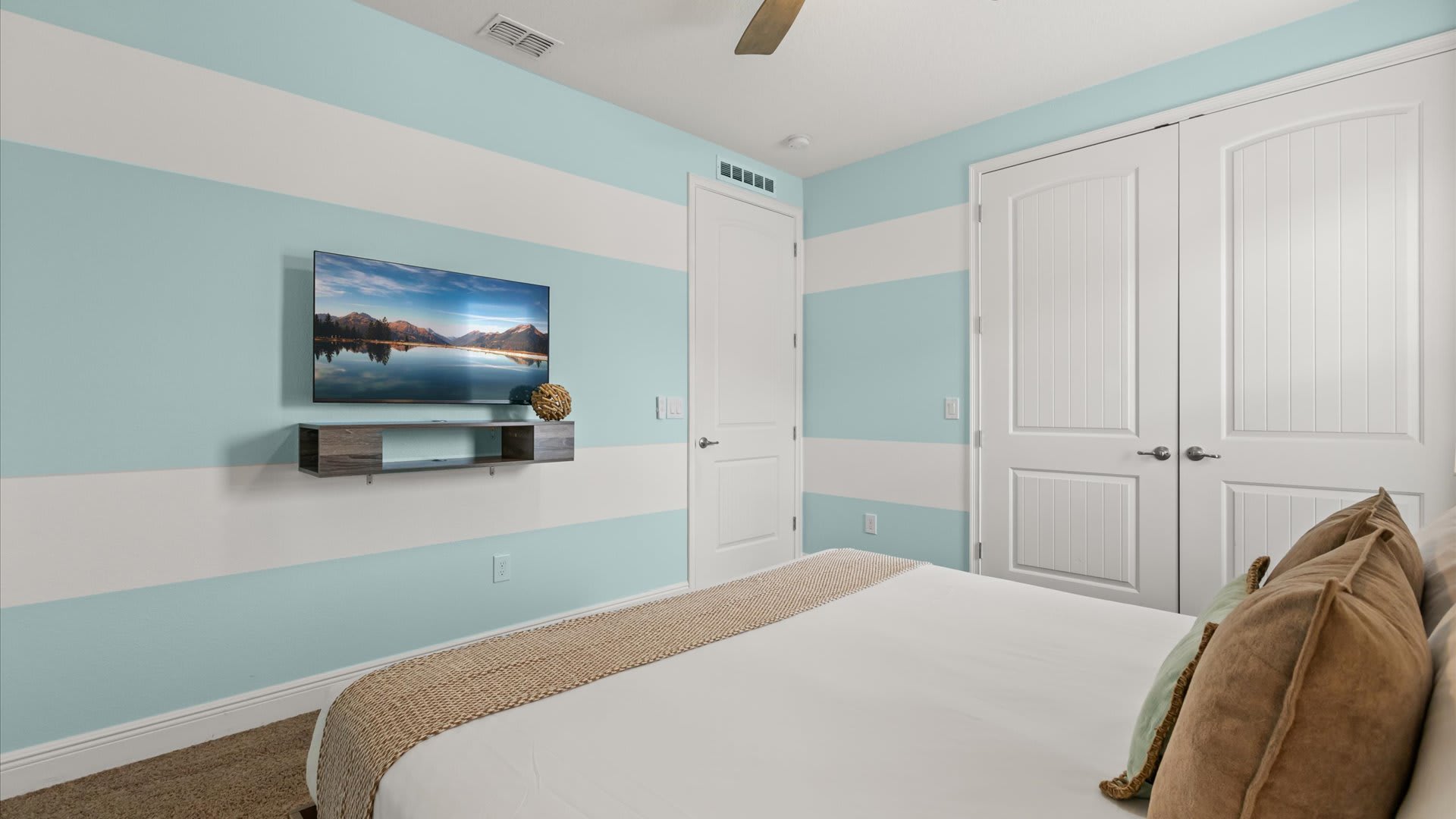 A bright bedroom with light blue and white striped walls, featuring a wall-mounted TV above a floating wooden shelf, and a bed with white bedding and a beige throw.