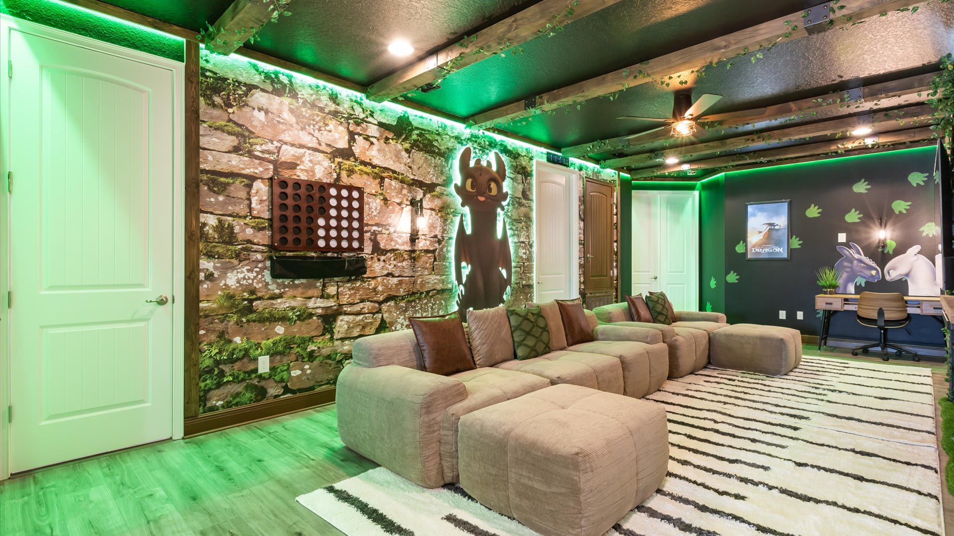 Themed game room with a large sectional sofa, a stone-look accent wall with a "How to Train Your Dragon" character cutout, and green LED lighting. The ceiling features dark beams with artificial vines, and the floor has a striped rug.