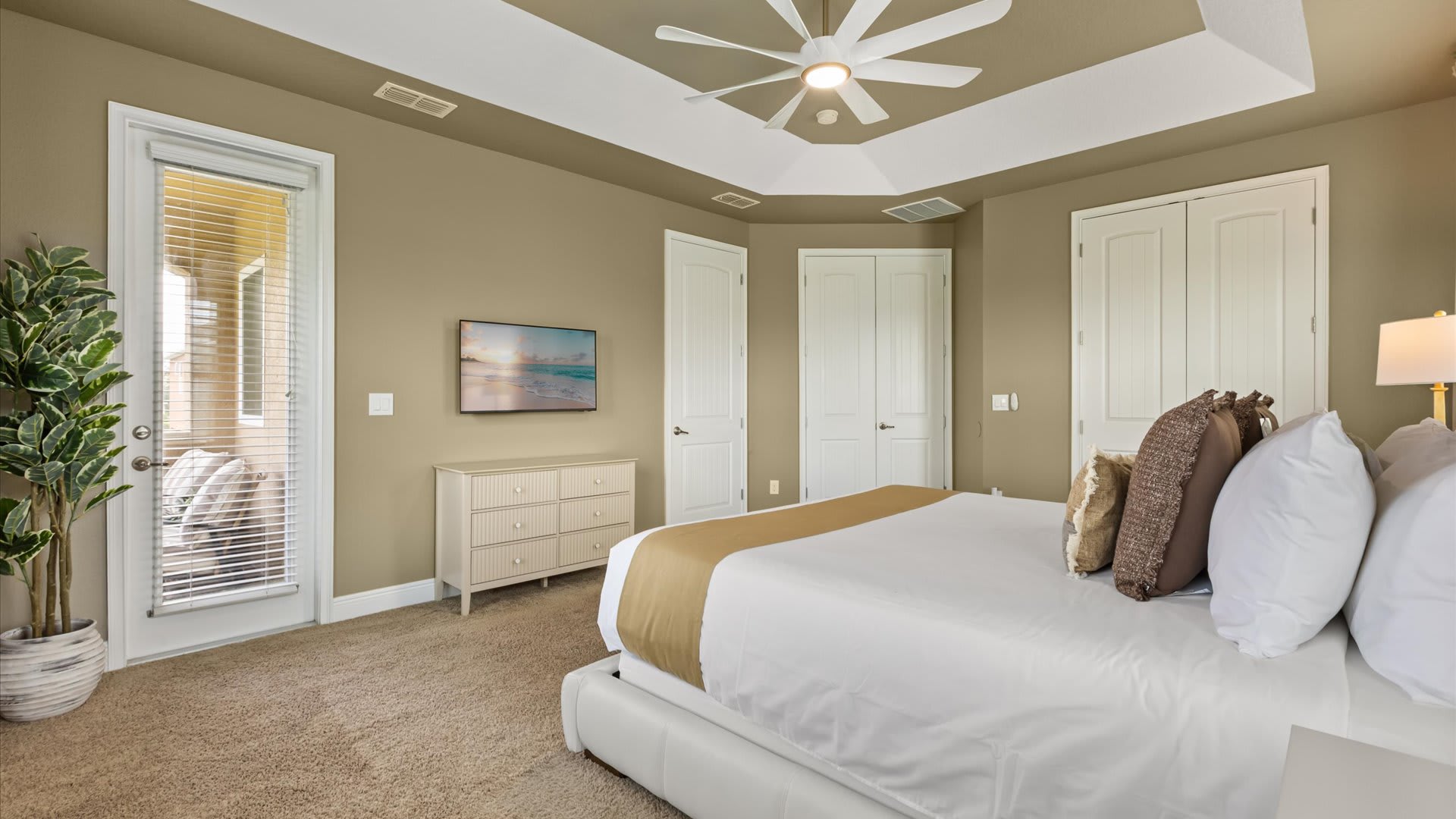Another view of the bedroom, showing a wall-mounted TV above a light wood dresser and a glass door leading to a balcony or patio. The room has a tray ceiling, ceiling fan, and carpeted floor.