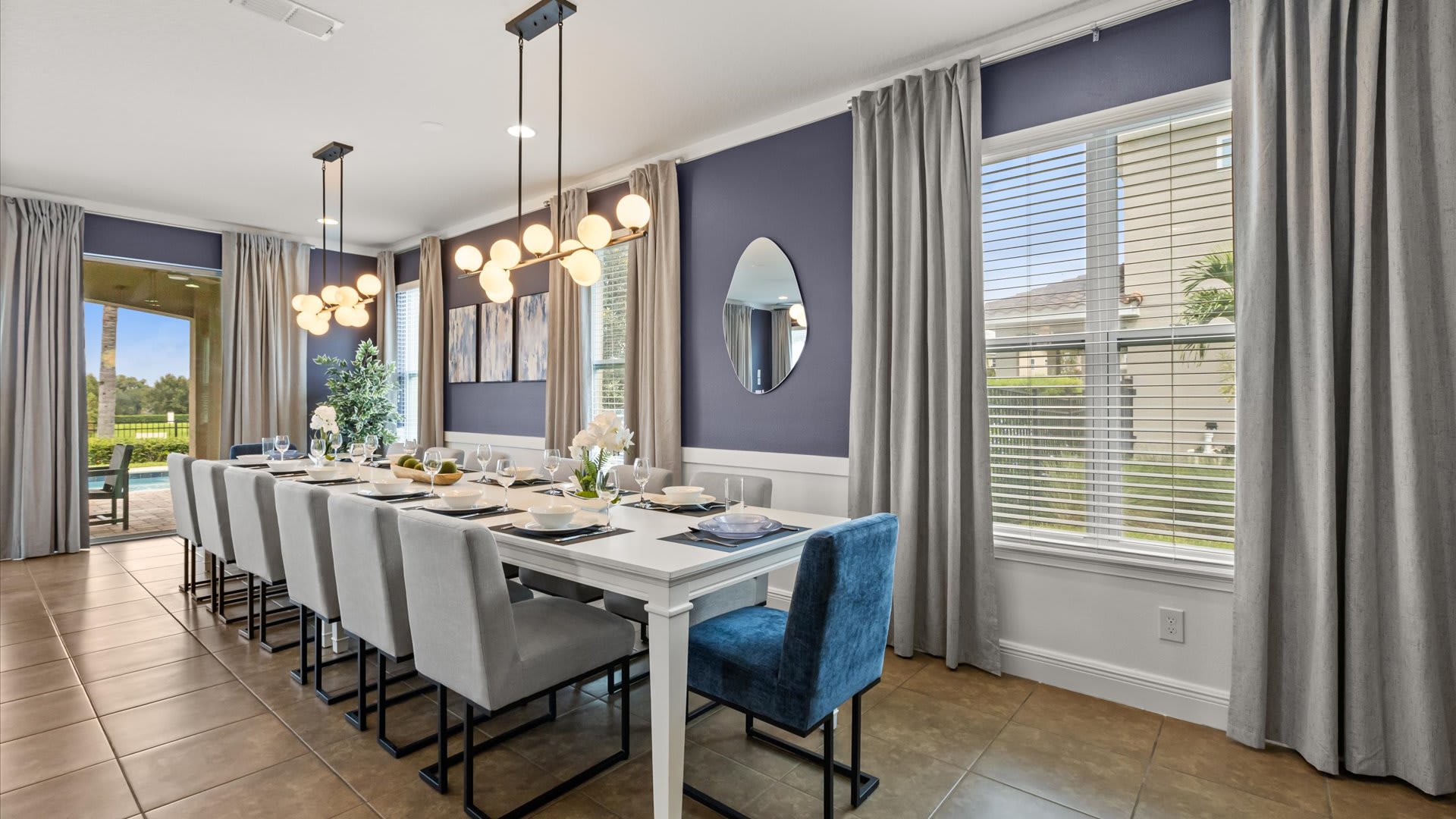 A spacious dining room features a long white dining table with seating for twelve, including one blue velvet chair, under two modern globe chandeliers. Large windows with blinds and grey curtains overlook the outdoor area.