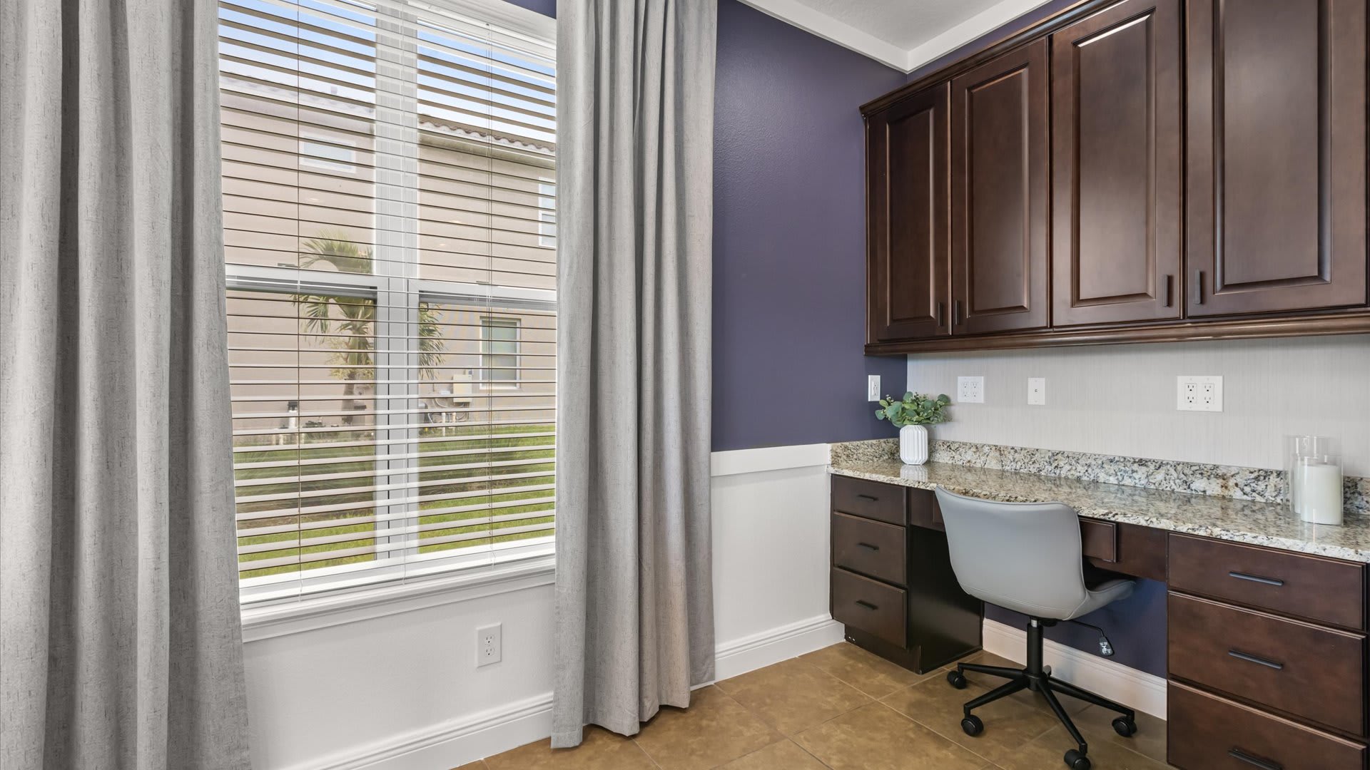 A dedicated office space with deep purple walls and white wainscoting, featuring a built-in dark wood desk with a granite countertop and matching upper cabinets. A large window with blinds and grey curtains overlooks the exterior.