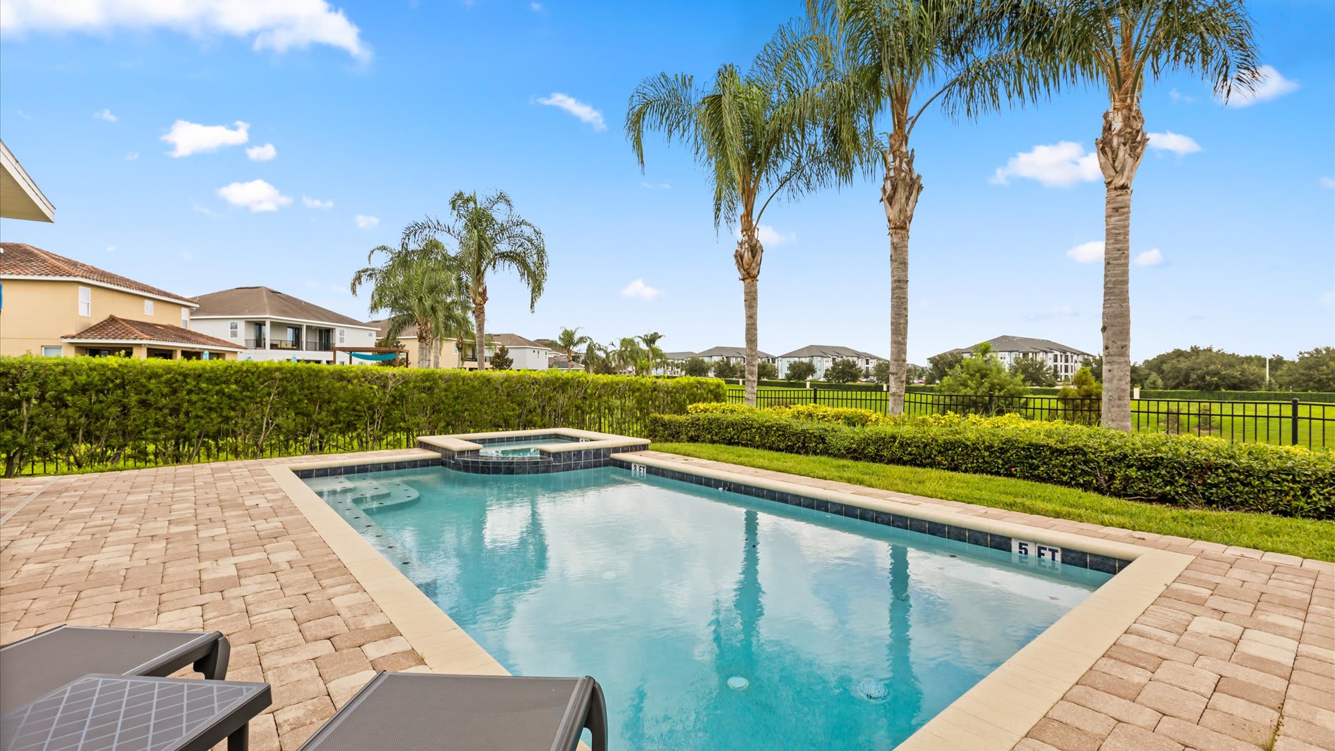 A private outdoor pool and integrated hot tub are set within a paver patio, bordered by a manicured hedge and palm trees under a bright blue sky. The pool depth is marked at 5 feet.