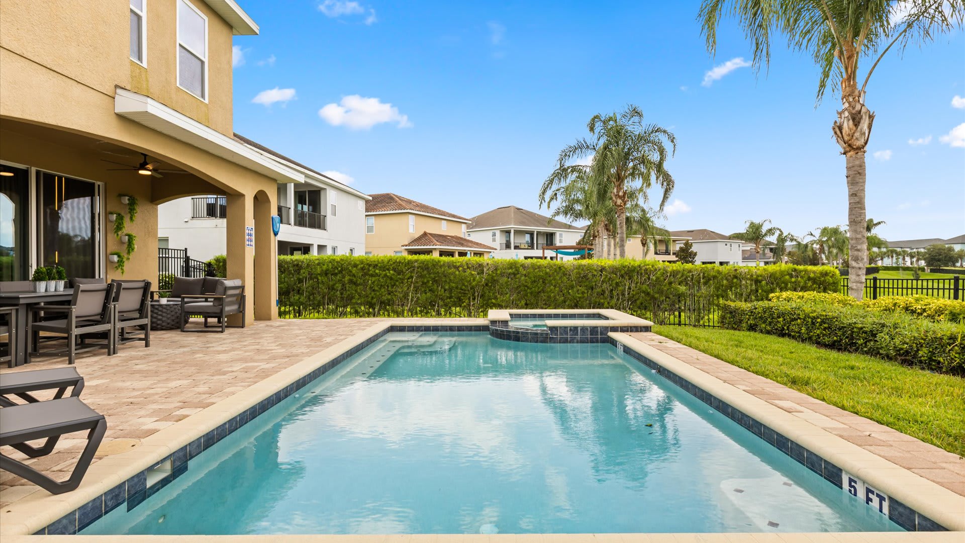 The exterior back of the house features a private pool and hot tub surrounded by a paver patio and lush green landscaping. A covered patio area with outdoor seating is visible to the left.