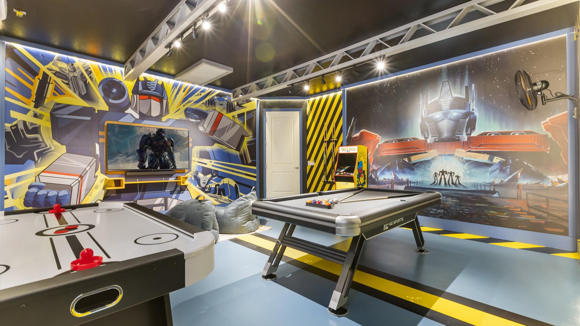 A vibrant game room features a Transformers theme with bold yellow and blue wall murals, an air hockey table, a pool table, and an arcade machine. Bright track lighting illuminates the space.
