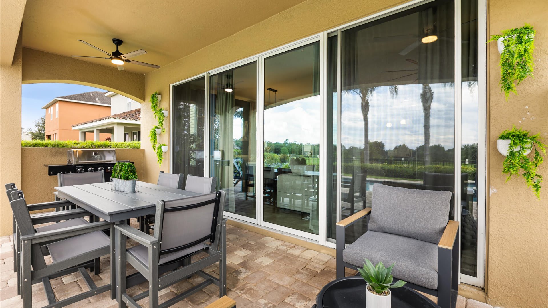 A covered outdoor patio featuring a grey dining table with six chairs and a separate lounge chair, all on a paved surface. Large sliding glass doors lead into the house, and hanging plants adorn the walls.